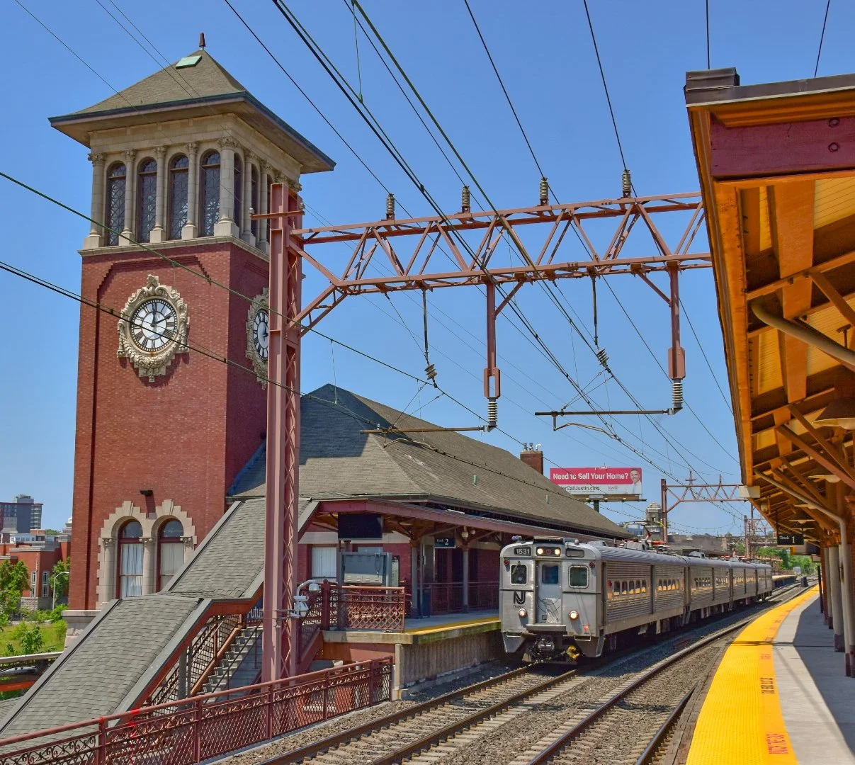 A silver train arriving at a station with a historic brick clock tower and platform on a sunny day.