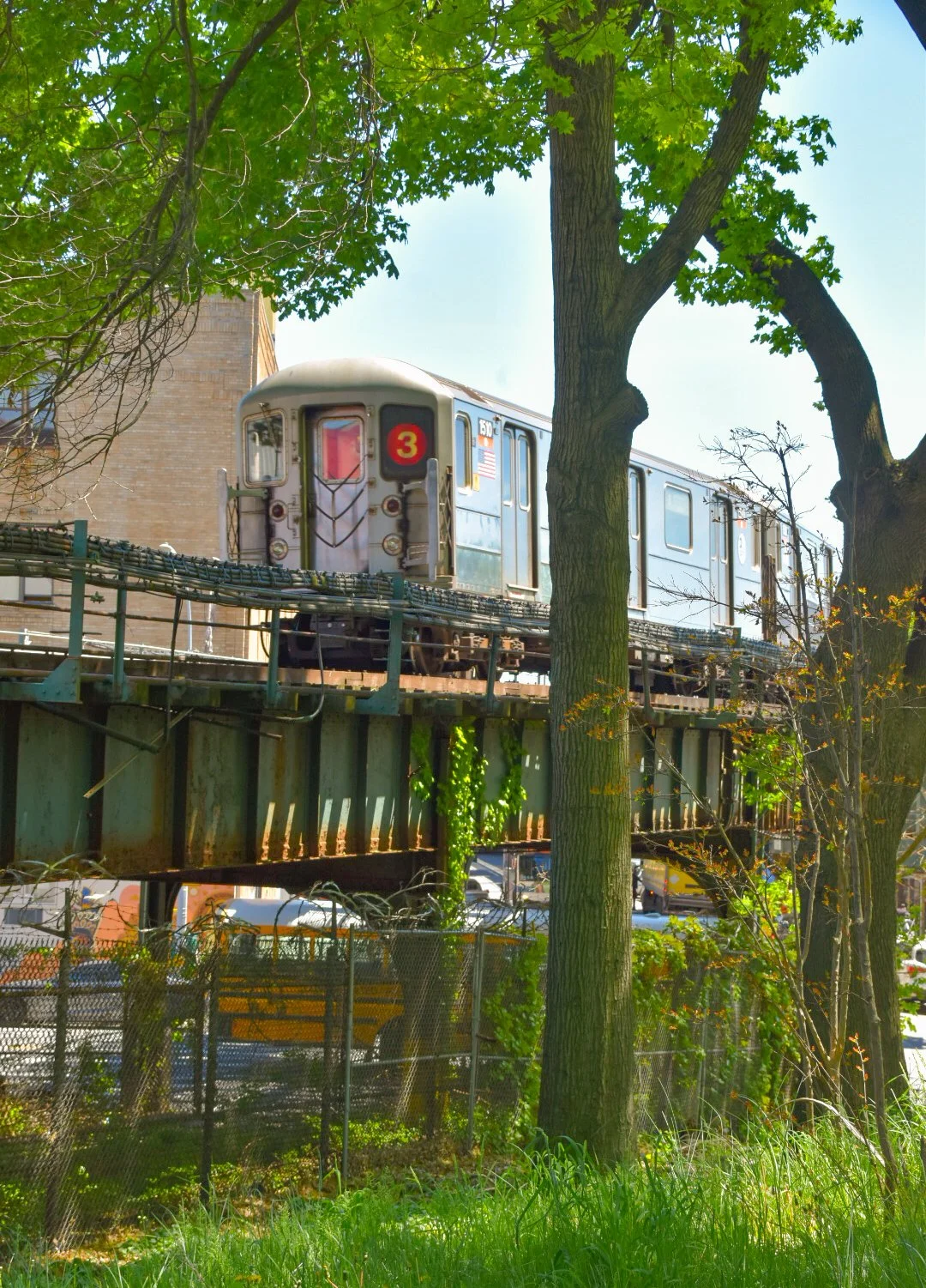 An elevated train on a track surrounded by trees and a fenced area, with a brick building in the background.
