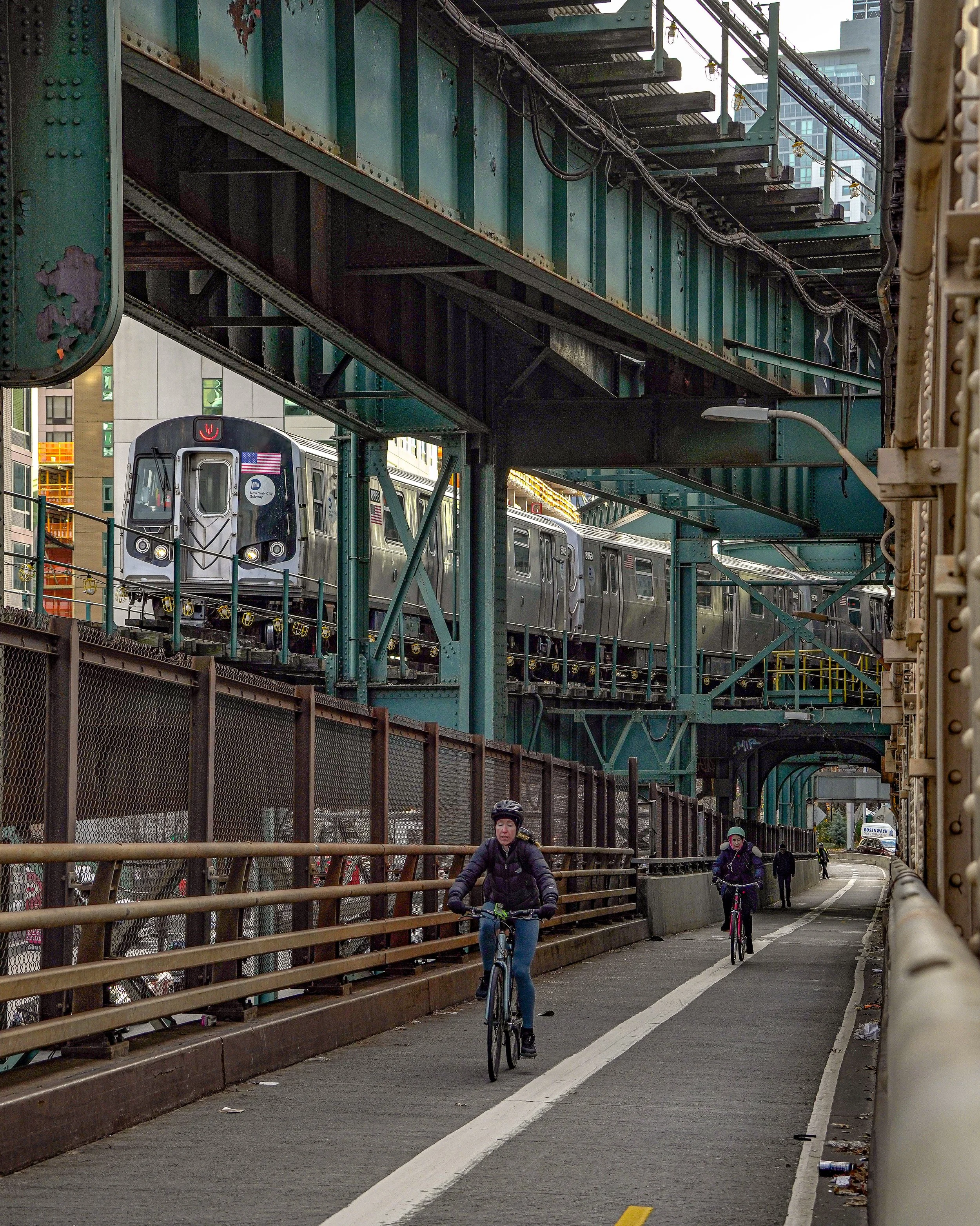 Bikers riding on a designated bike lane under an elevated train track with a train passing above in an urban area.