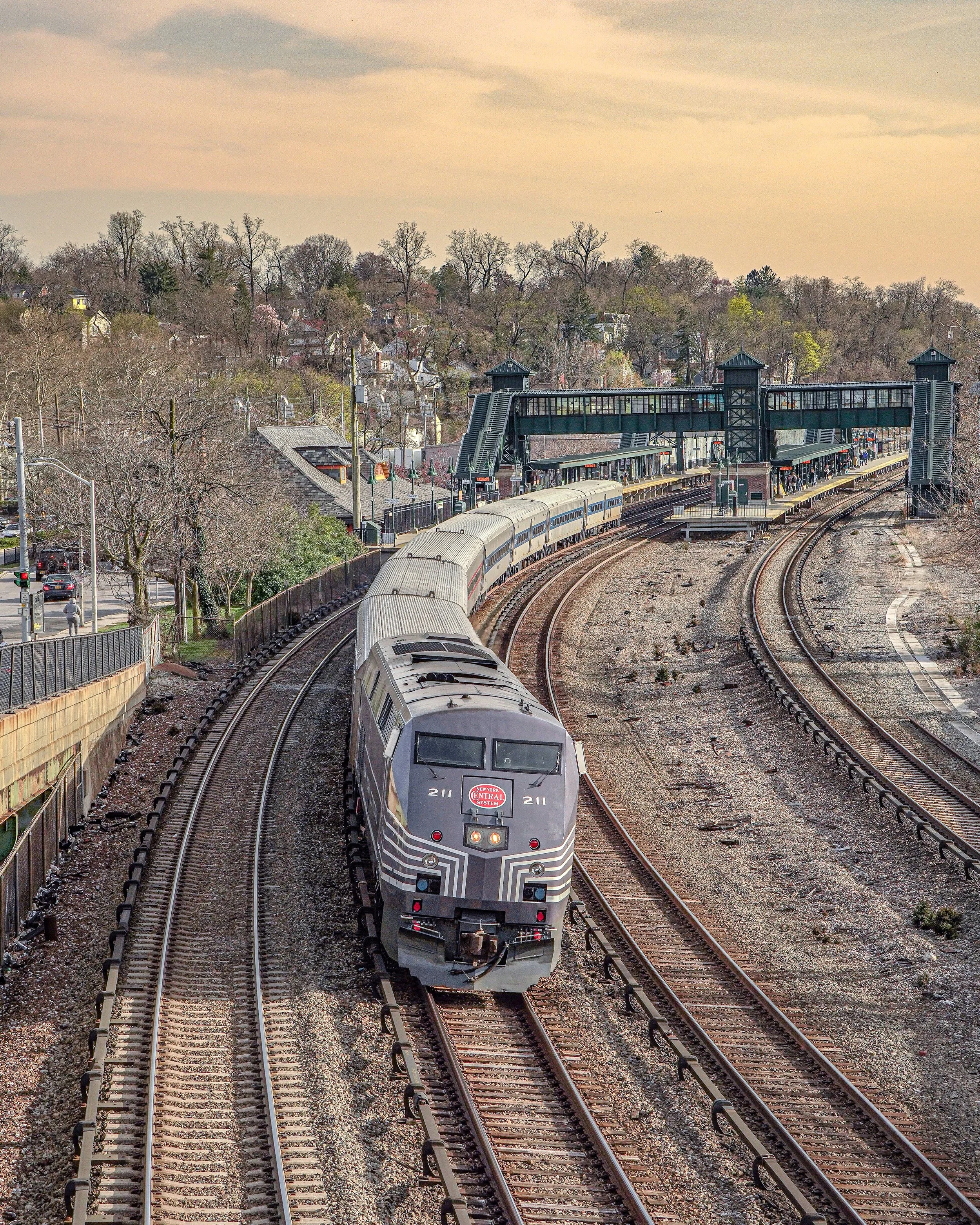 A black and gray commuter train traveling on curved tracks towards a city station with a green bridge and residential area with trees in the background.
