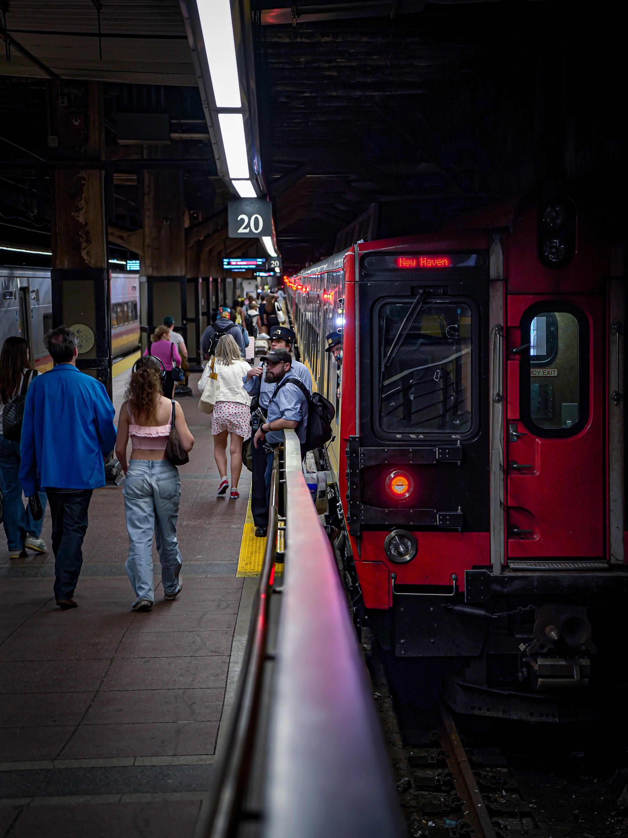 People waiting on a subway platform, with a red train labeled 'New Haven' waiting for departure, under a dark ceiling with lit signs and a speed limit of 20.