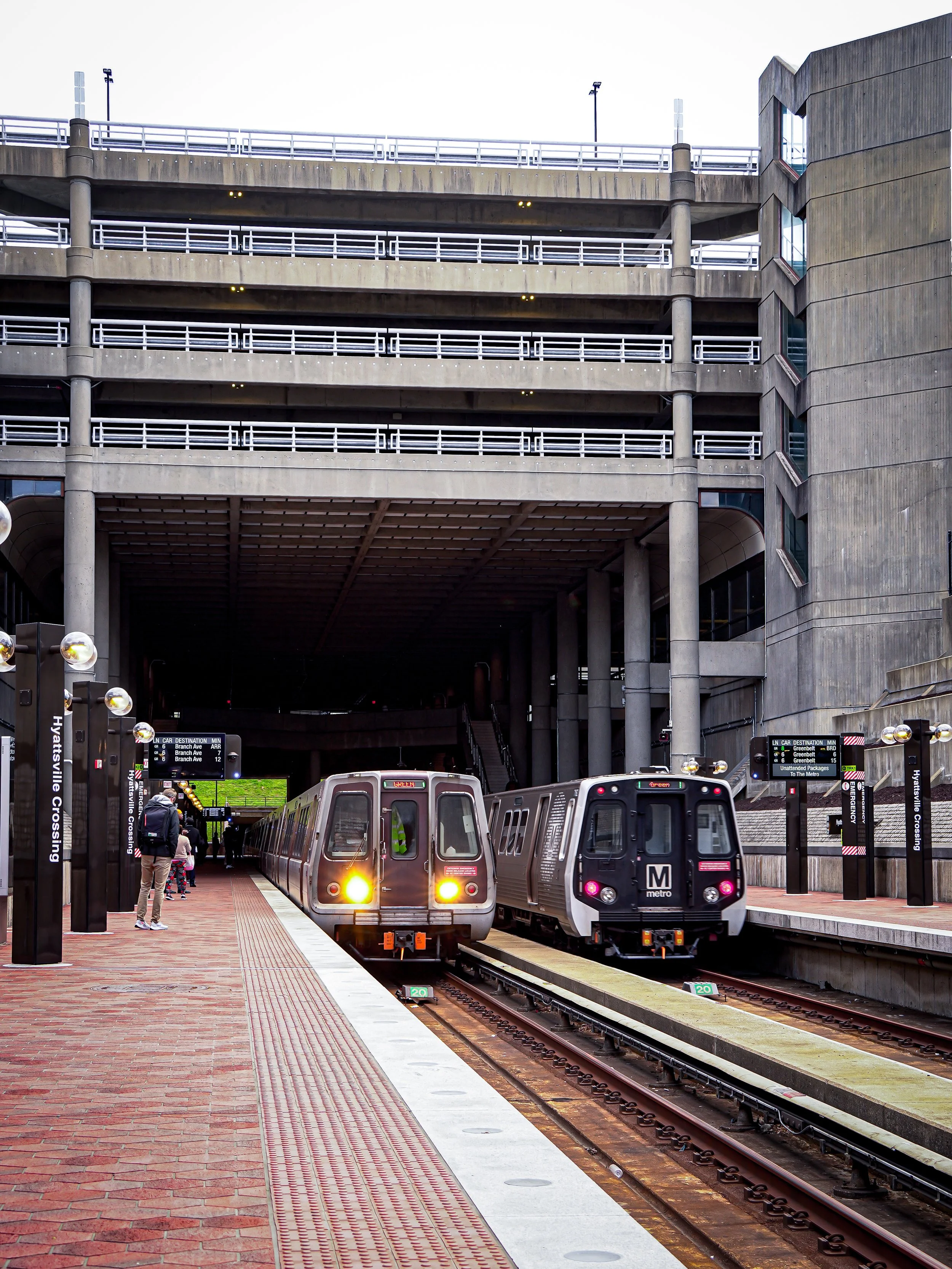 Two light rail trains at a platform at an urban transit station beneath a concrete parking garage structure.