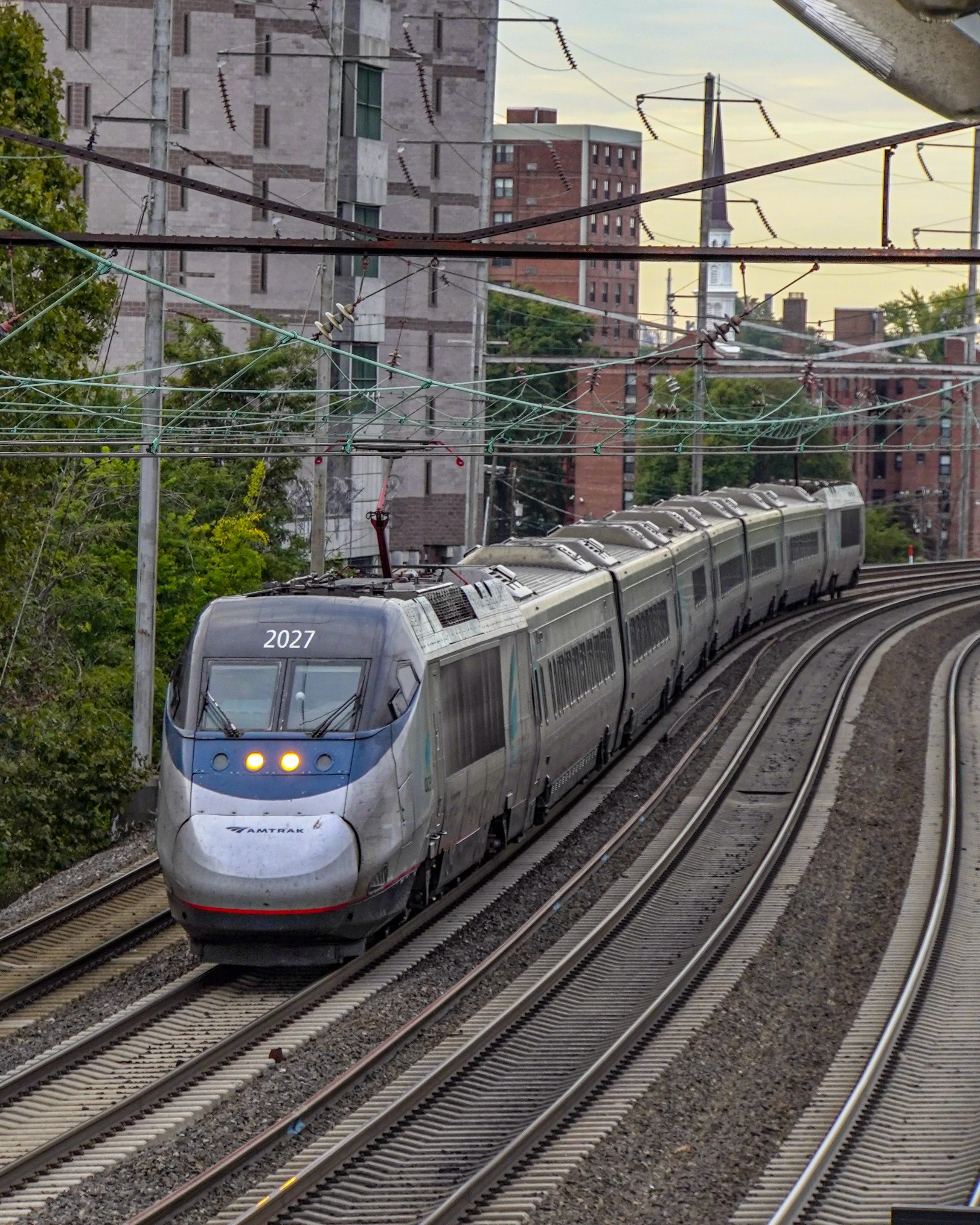A modern Amtrak passenger train traveling on elevated tracks through an urban area with buildings and a church steeple in the background.
