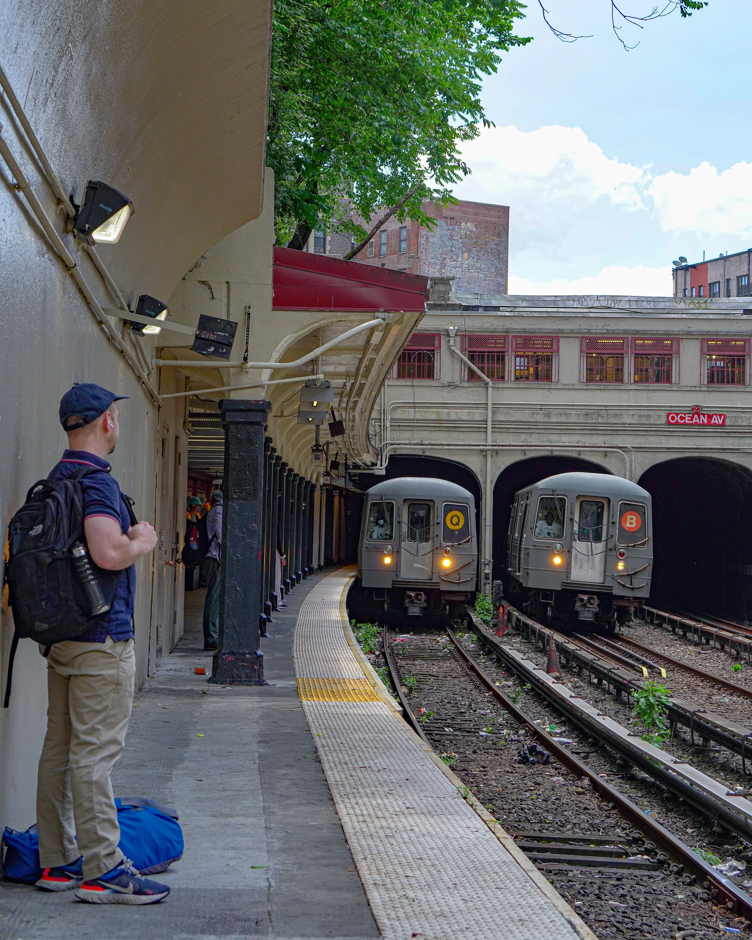 A subway station with two trains on parallel tracks, a man with a backpack standing on the platform, and a cityscape in the background.