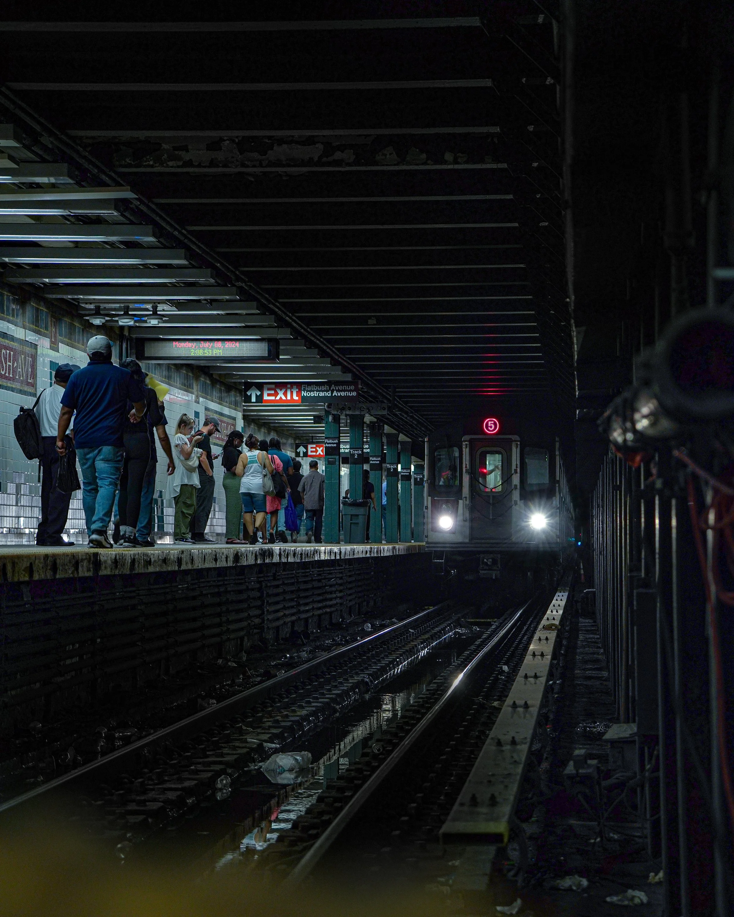 People standing on a subway platform as a train approaches in a dimly lit underground station.