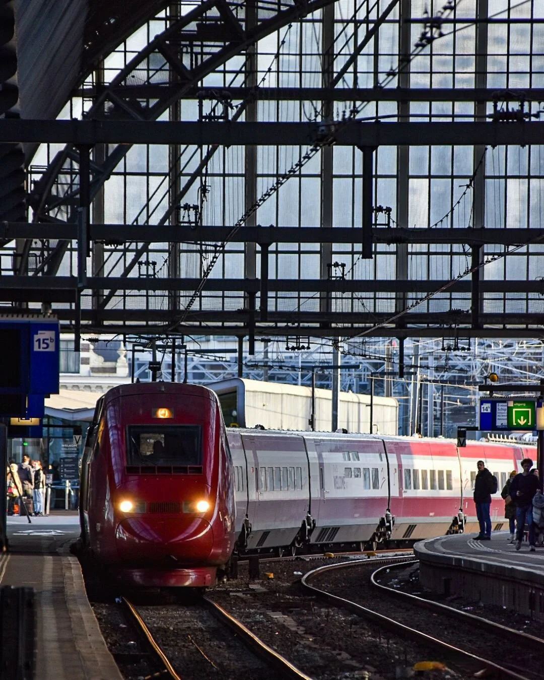 Red high-speed train arriving at a busy train station platform with passengers and overhead wiring.
