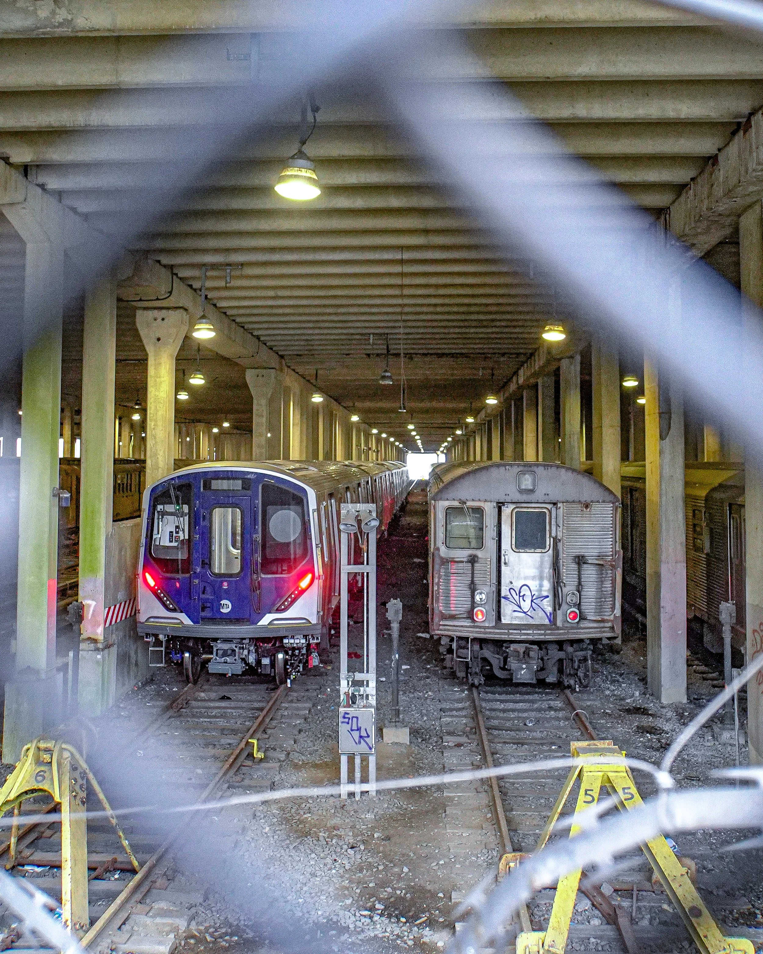 Inside a subway train yard with two trains parked on parallel tracks, viewed through a chain-link fence, with overhead lighting illuminating the scene.
