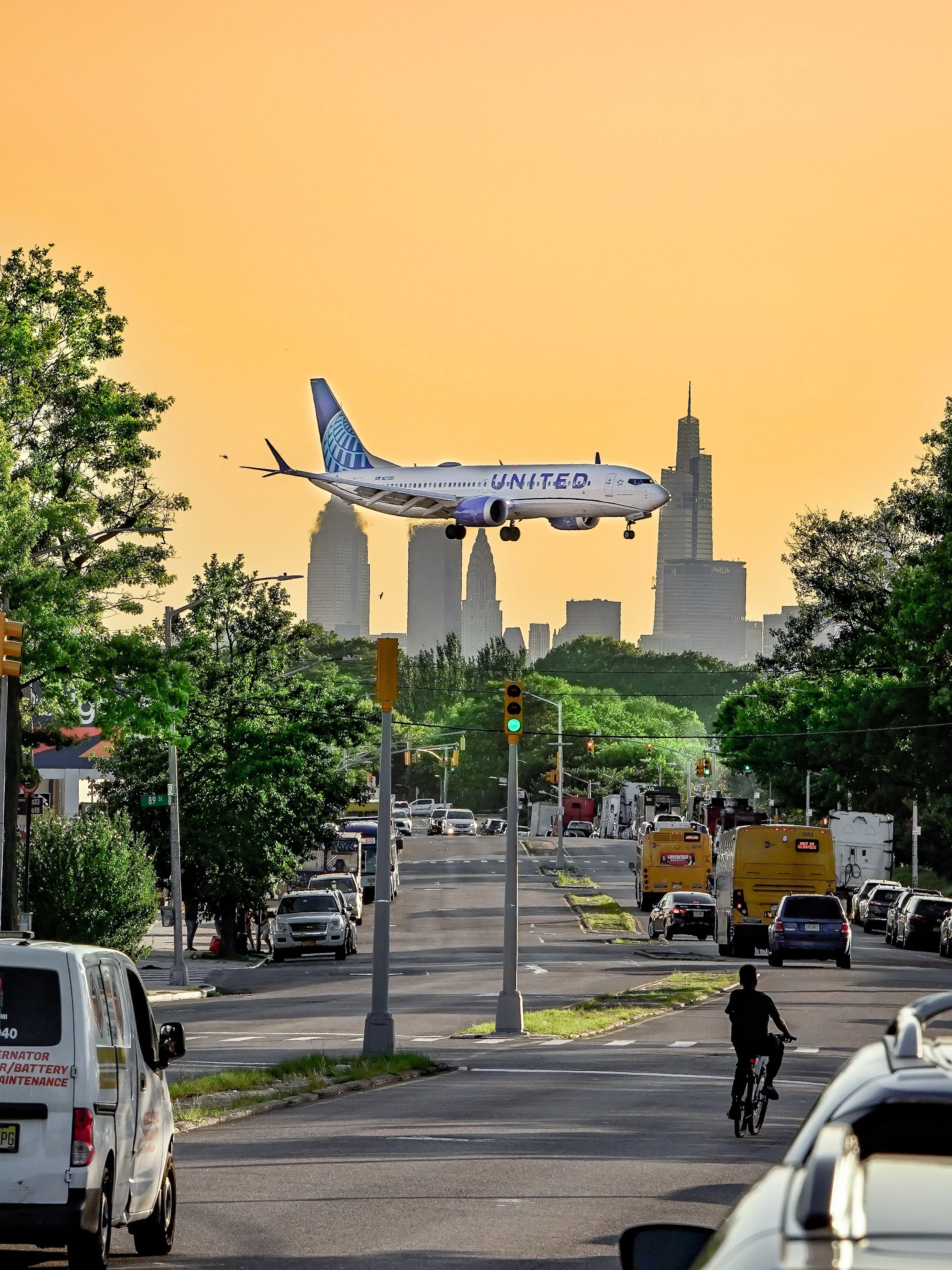 An airplane flying low over a city street with traffic and a cyclist, with the skyline in the background during sunset.