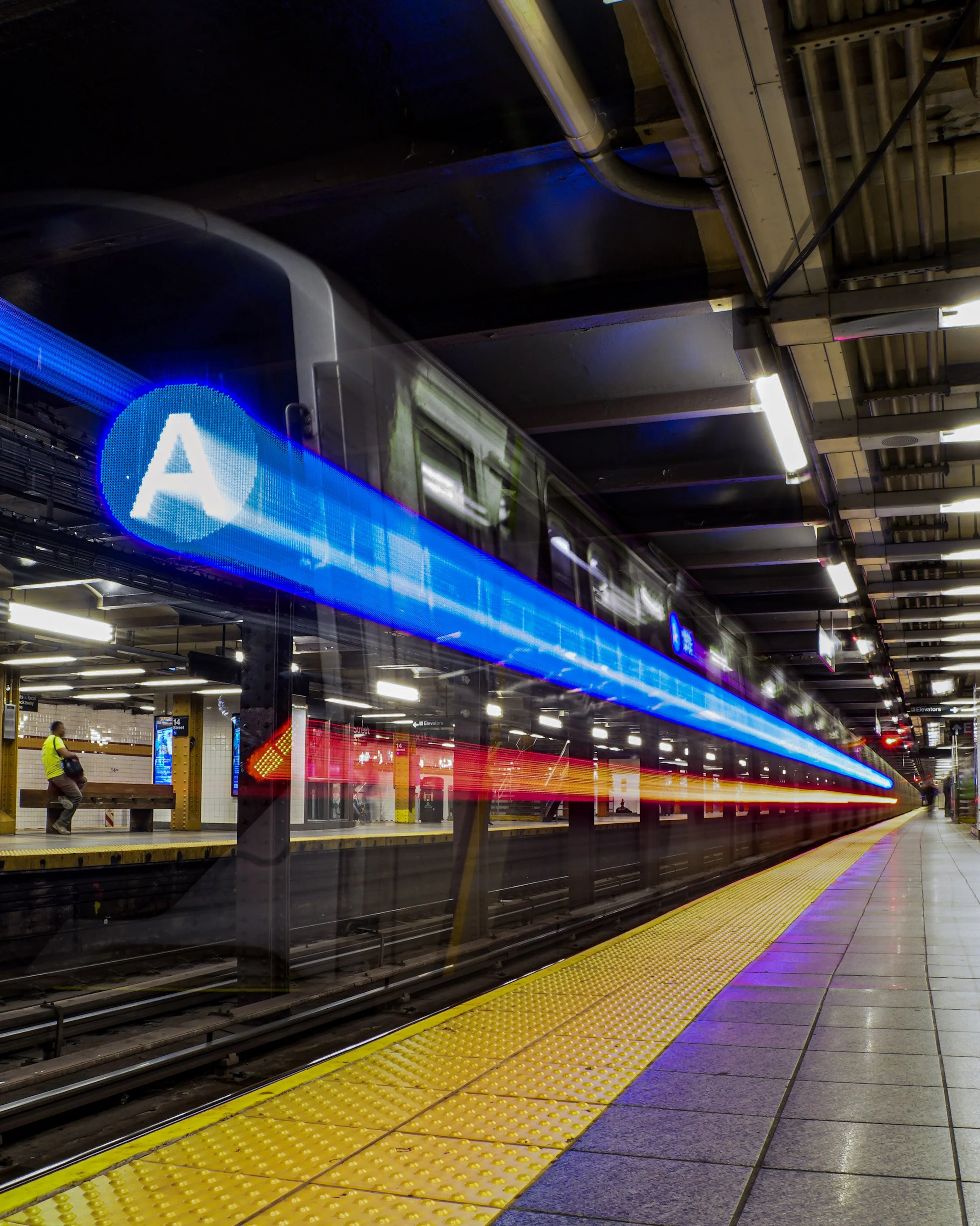 Long exposure photo of an A subway train departing at an underground station with blue and red light trails, yellow tactile paving, and a person sitting on a bench on the platform.