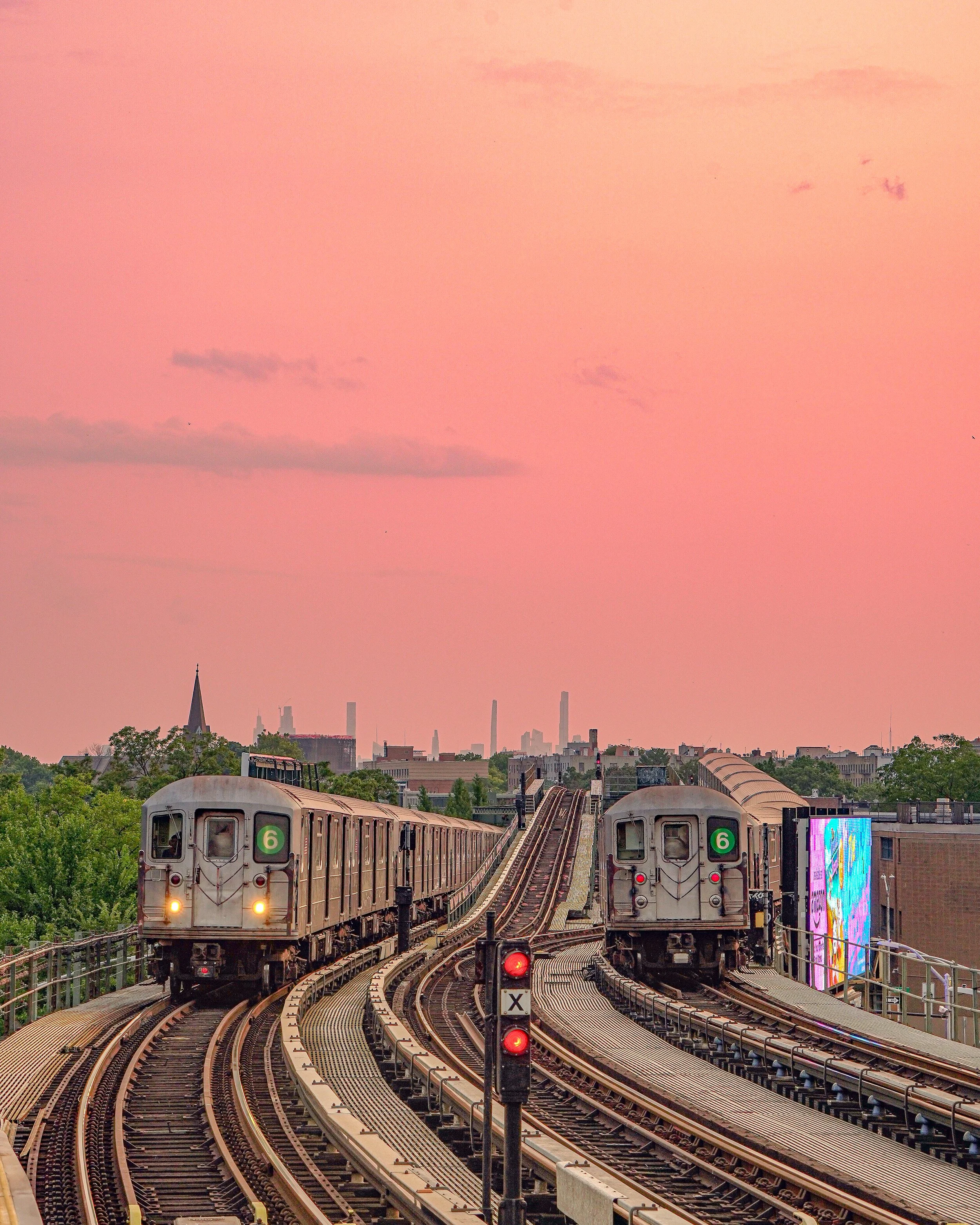 Two subway trains on elevated tracks against a pink sunset sky with city skyline in the background.