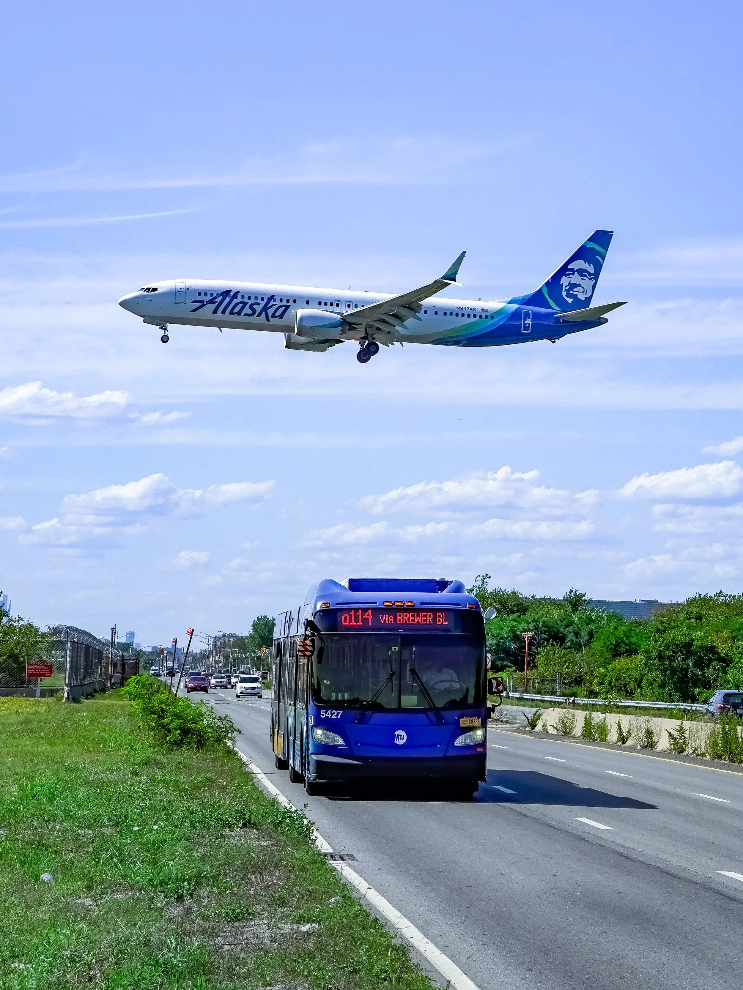 An Alaska Airlines airplane flying low in the sky above a city road, with a blue bus driving on the street below on a sunny day.