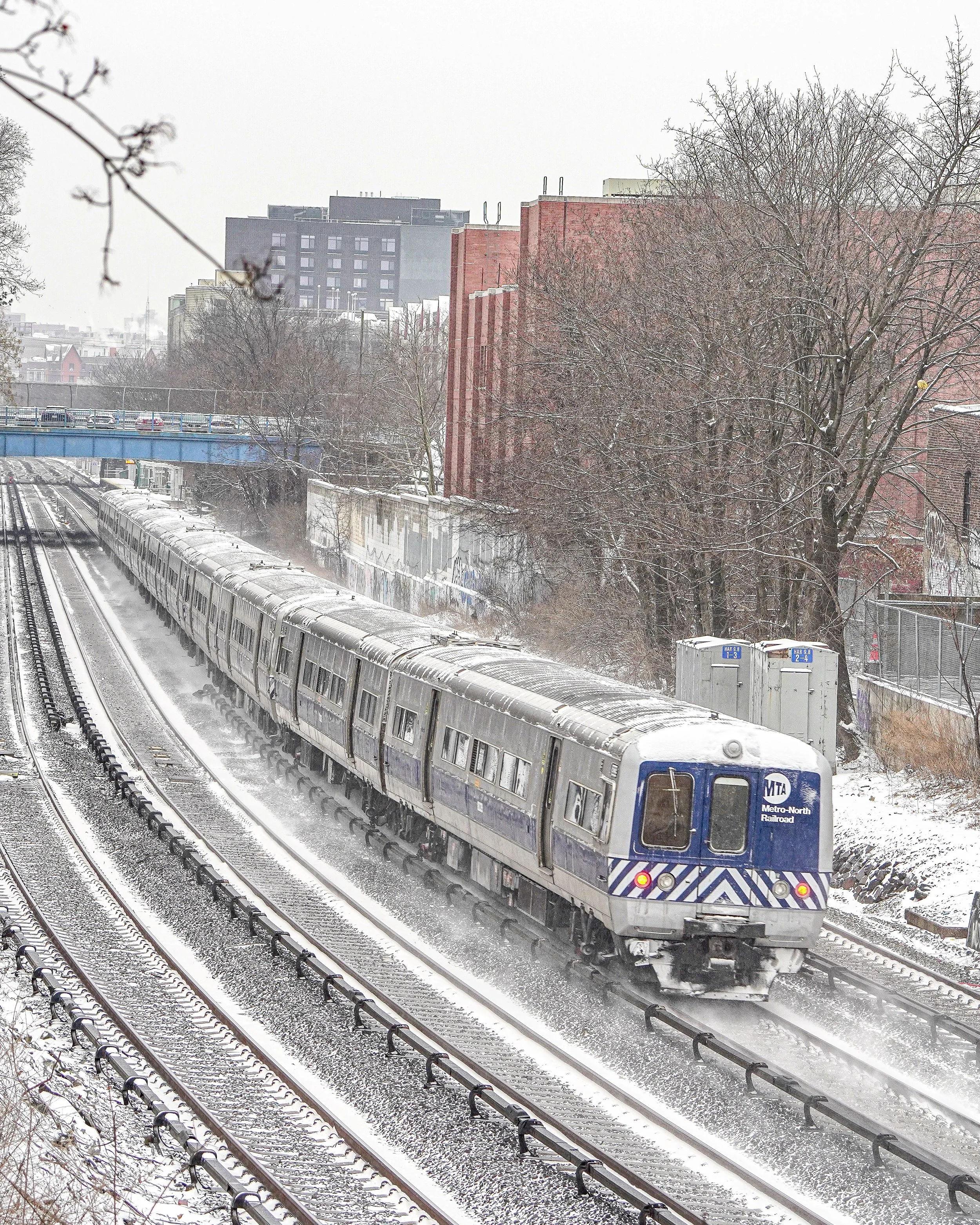 A Metro-North train traveling on snow-covered railway tracks through an urban area with trees and buildings in the background.