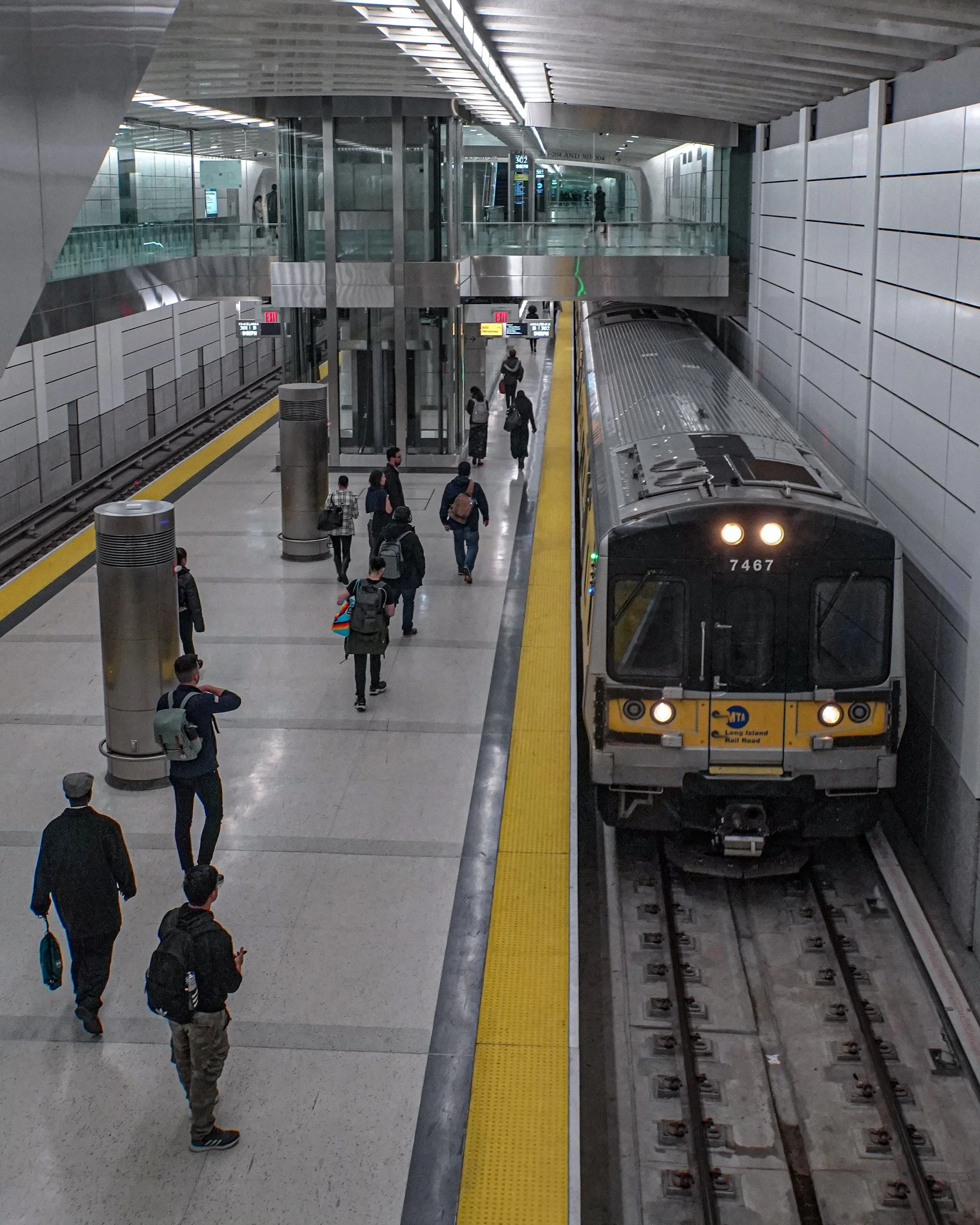 People walking on a subway station platform with an arriving subway train on the right. The station has modern design with glass and metal fixtures.