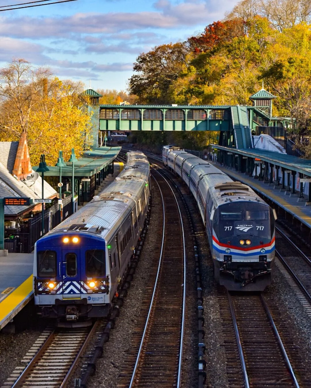 Two trains approaching a station with a green bridge overhead, surrounded by autumn-colored trees on a clear day.