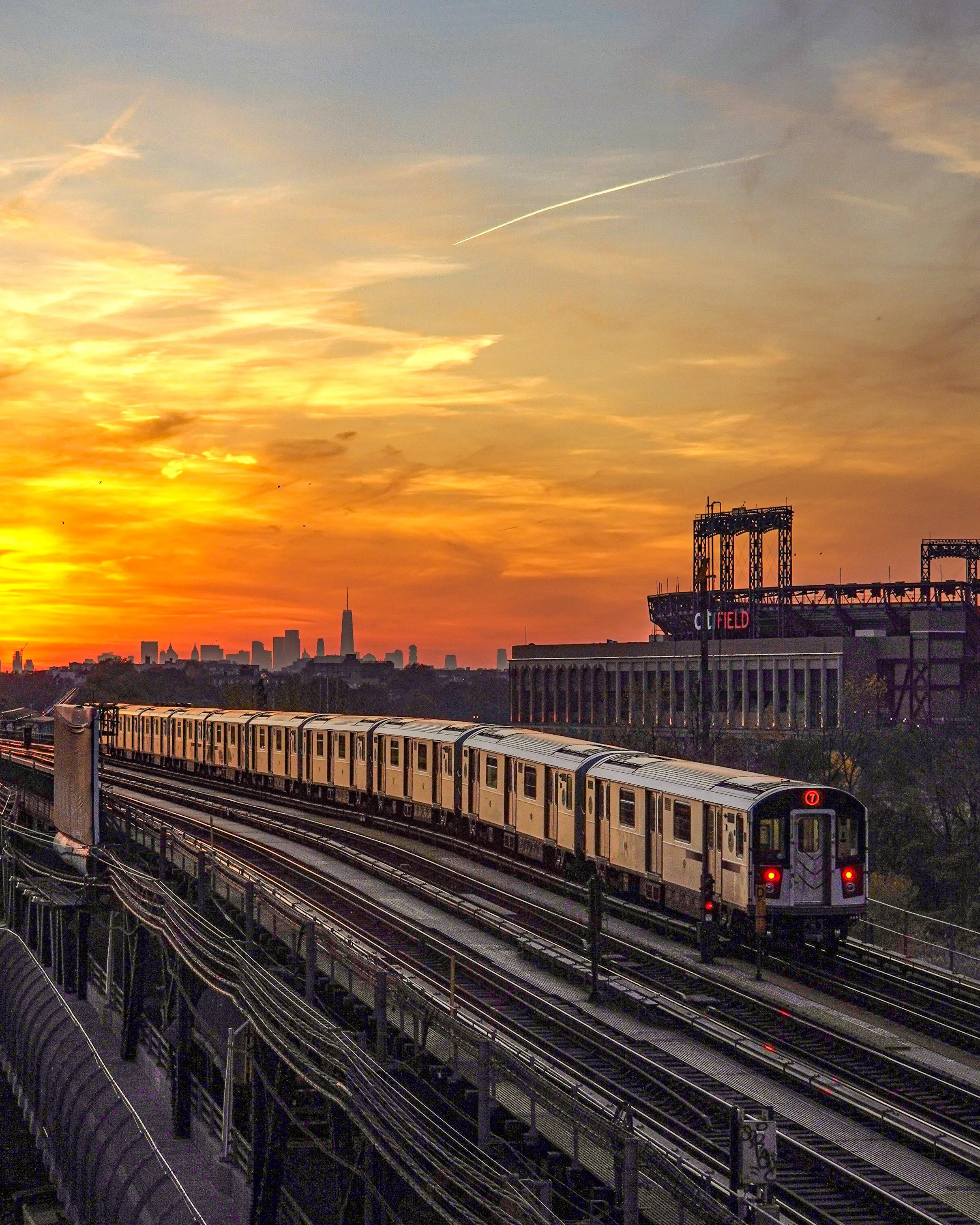 A subway train traveling on elevated tracks during sunset, with a city skyline in the background.