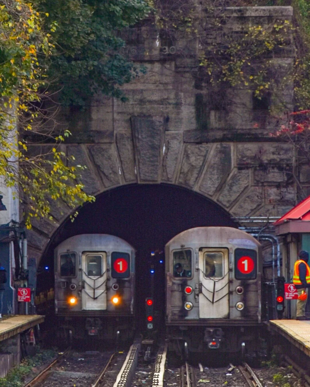 Two subway trains emerge from a dark tunnel in an urban setting, with a stone arch above, surrounded by trees and some station structures to the sides.