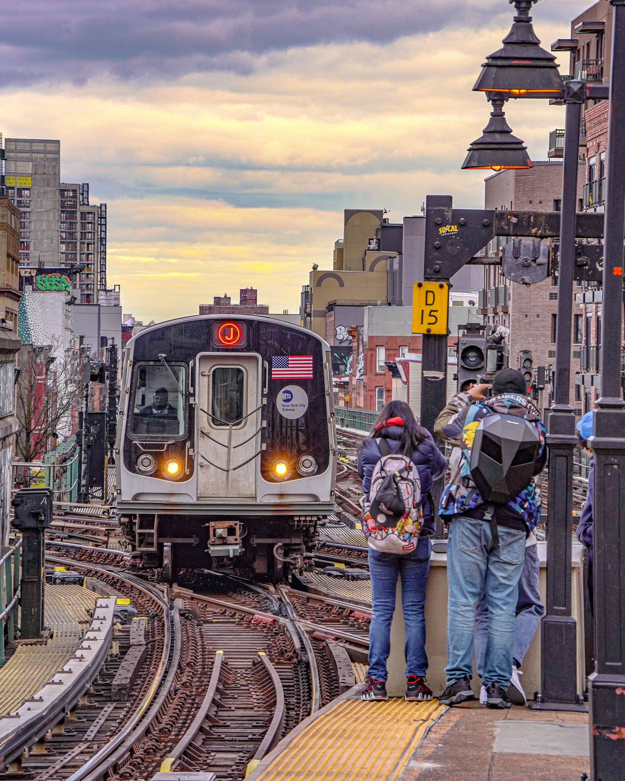 A New York City subway train arriving at a station with three pedestrians on the platform, two women and one man, during dusk with a cloudy sky.