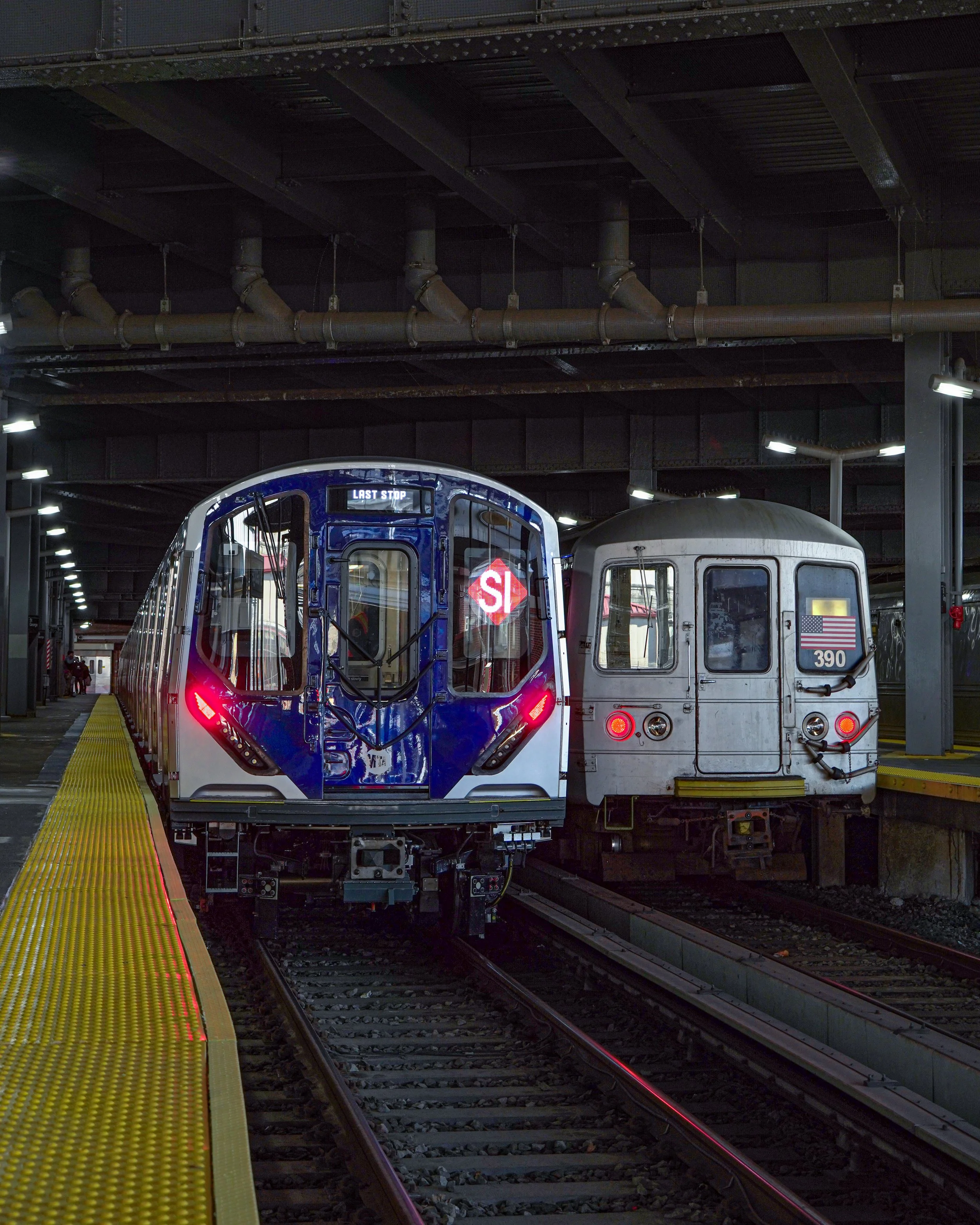 Two subway trains parked side by side on underground tracks, one modern blue and white, the other older gray, with overhead pipes and ceiling lights visible.