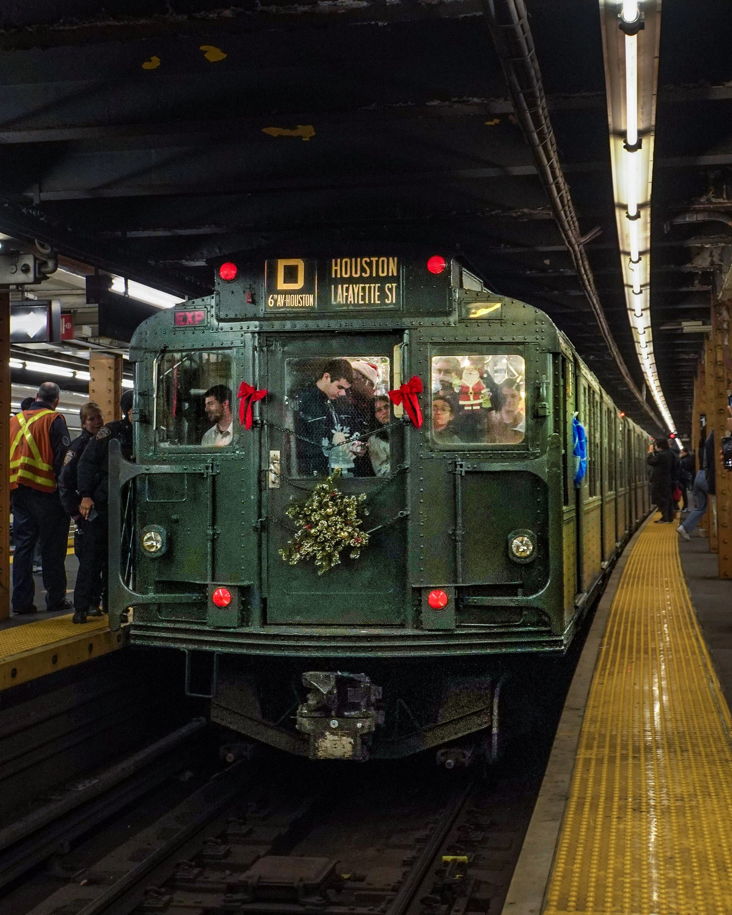 A vintage green train decorated with Christmas ornaments and bows is arriving at a station, with passengers and station staff standing on the platform.