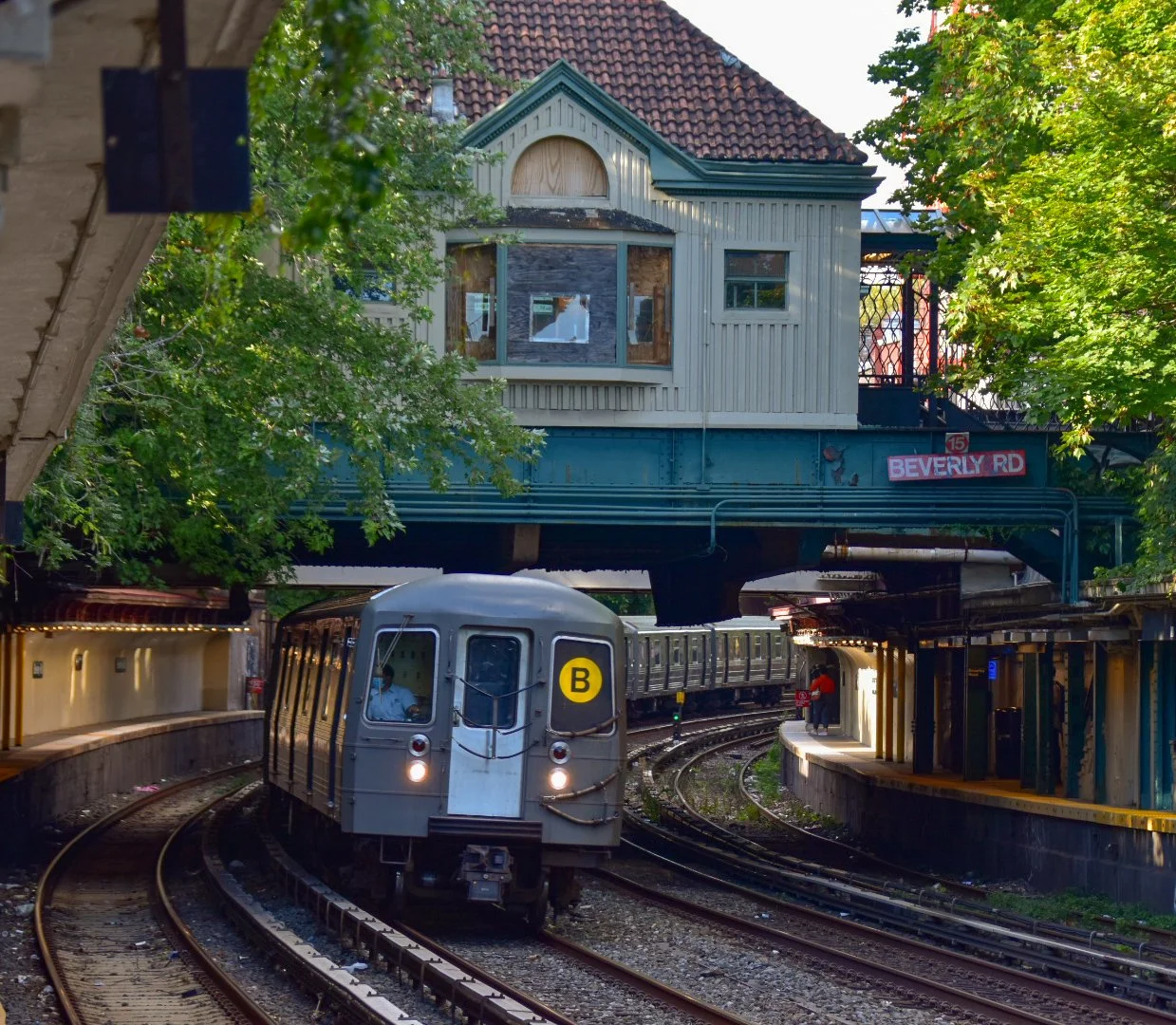 An elevated train passing through a neighborhood with trees, under a building with a red tiled roof and a sign that says Beverly Rd, under the tracks.