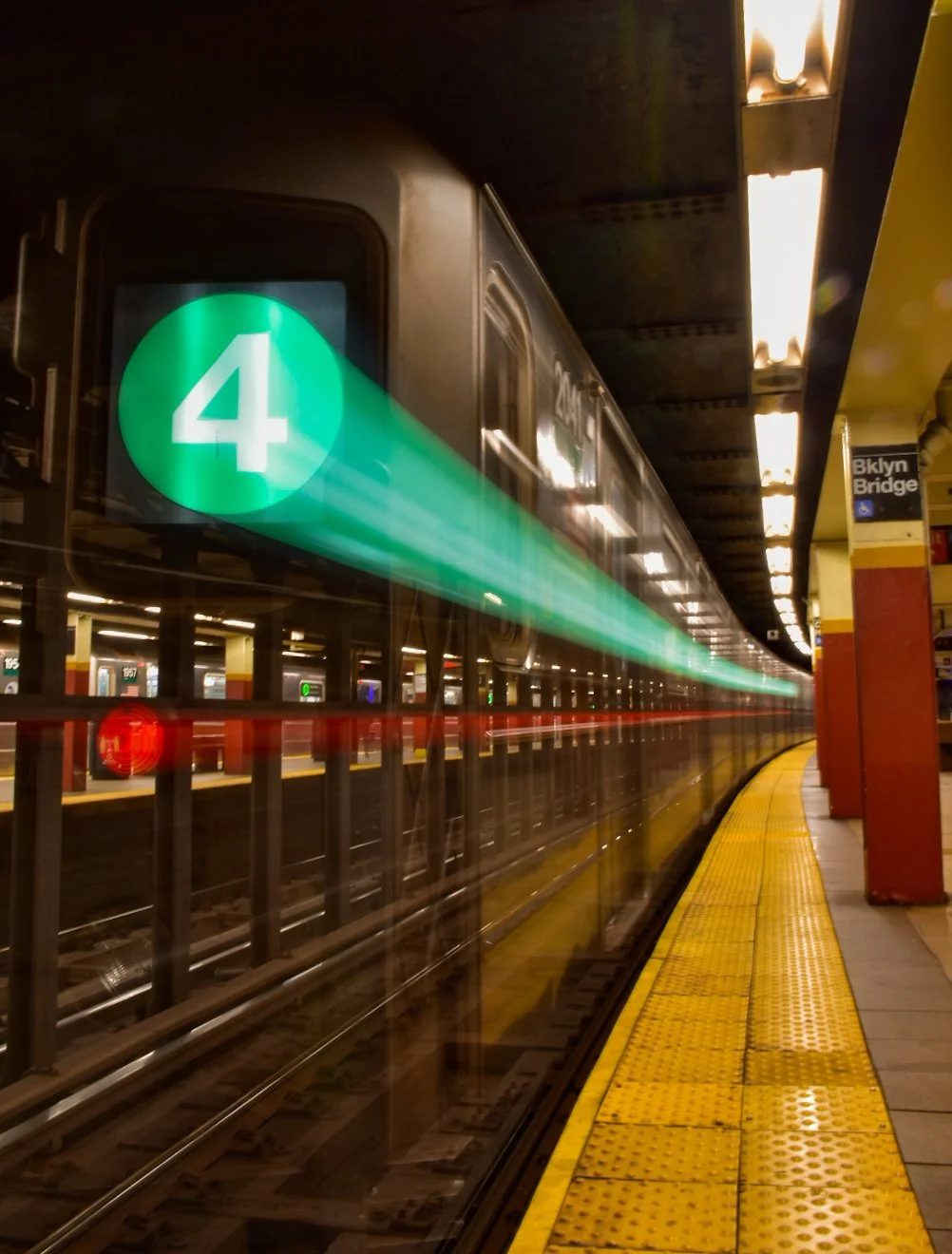 Subway train at a station platform with a green number 4 sign indicating the train route, Brooklyn Bridge signage visible, and fluorescent lights overhead.