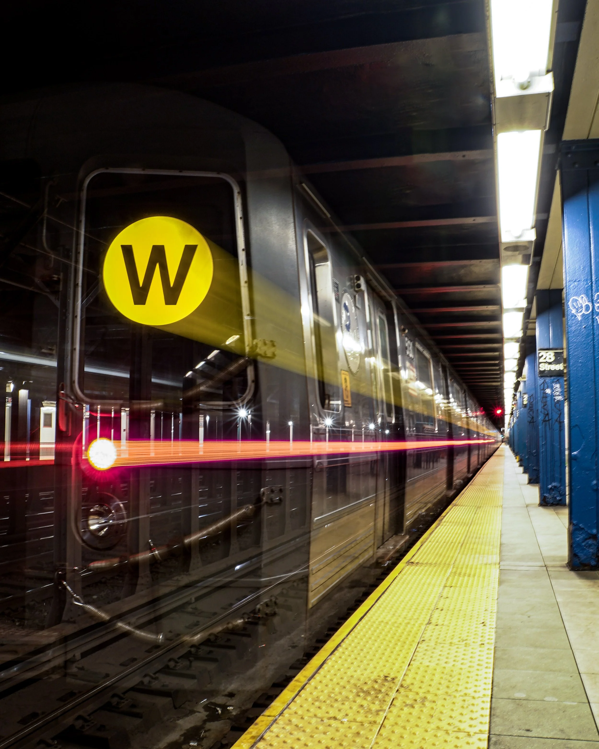 A subway train at a station platform with a yellow circle and a black 'W' sign. The photo is taken at night, with motion blur of the train showing red and white light streaks.