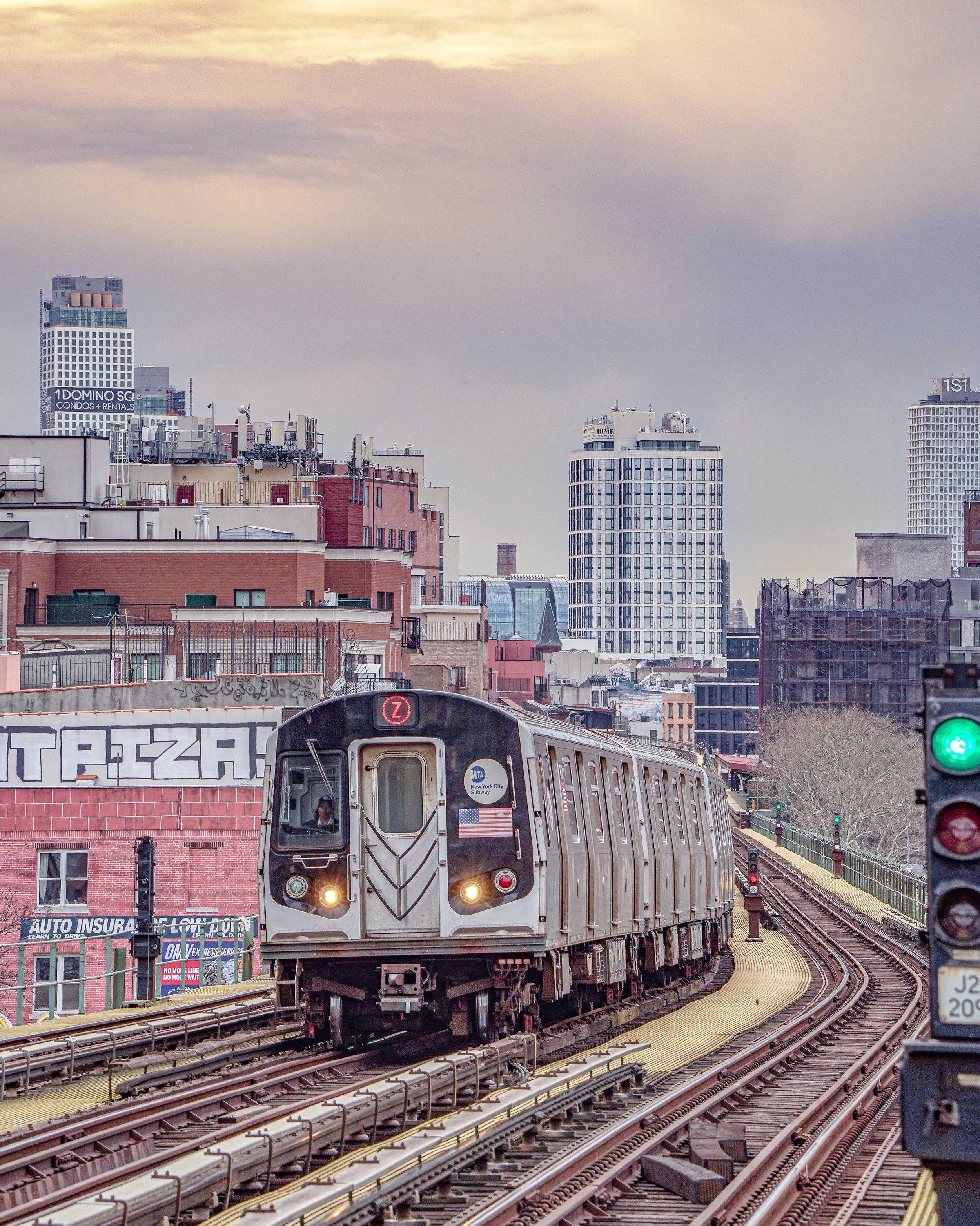 A subway train traveling on elevated tracks in an urban cityscape with tall buildings and graffiti on a building in the background.