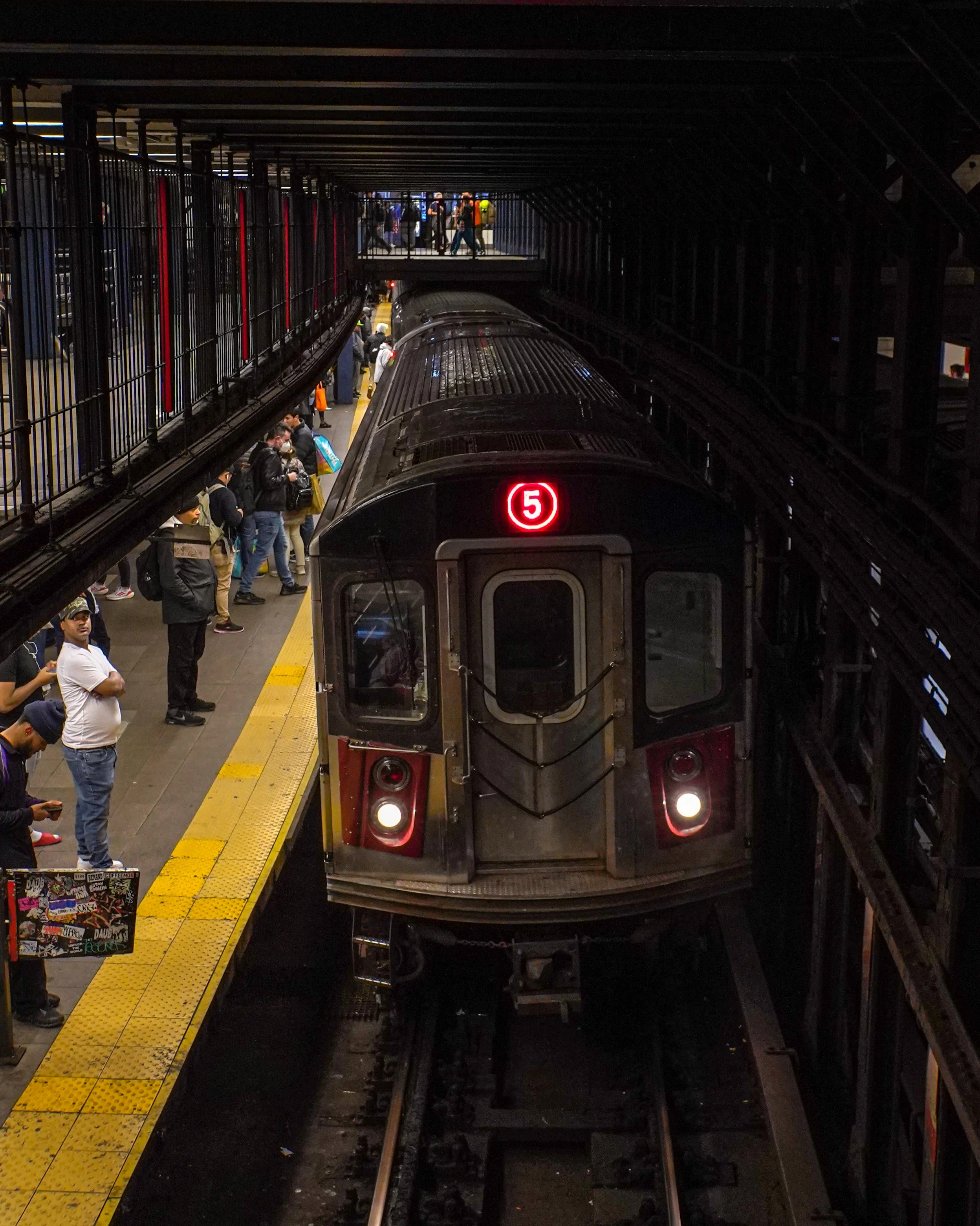 A subway train at a station platform with passengers waiting, illuminated by station lights, and a sign displaying the number 5.