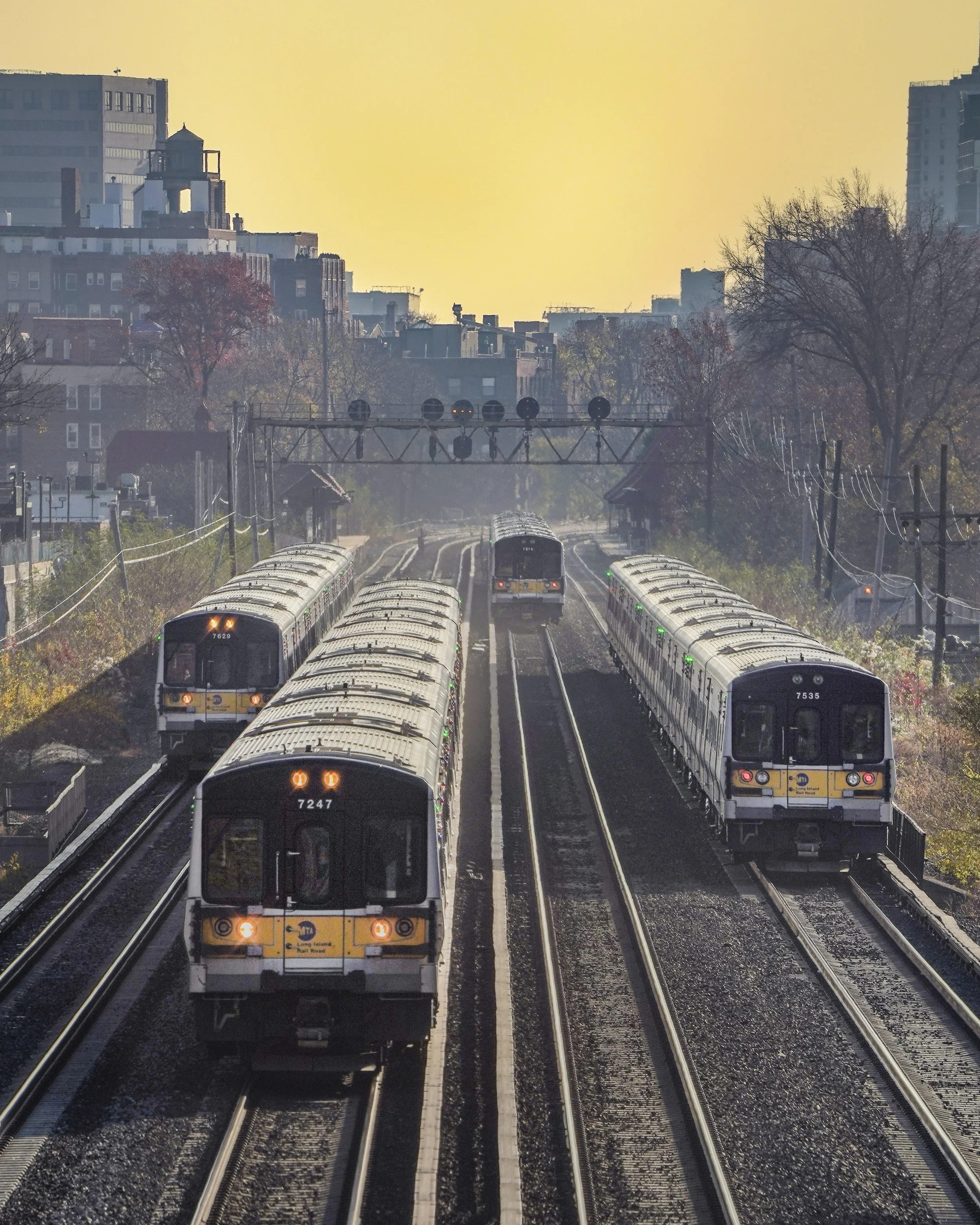 Multiple subway trains moving along railroad tracks with urban buildings in the background during sunset or sunrise.
