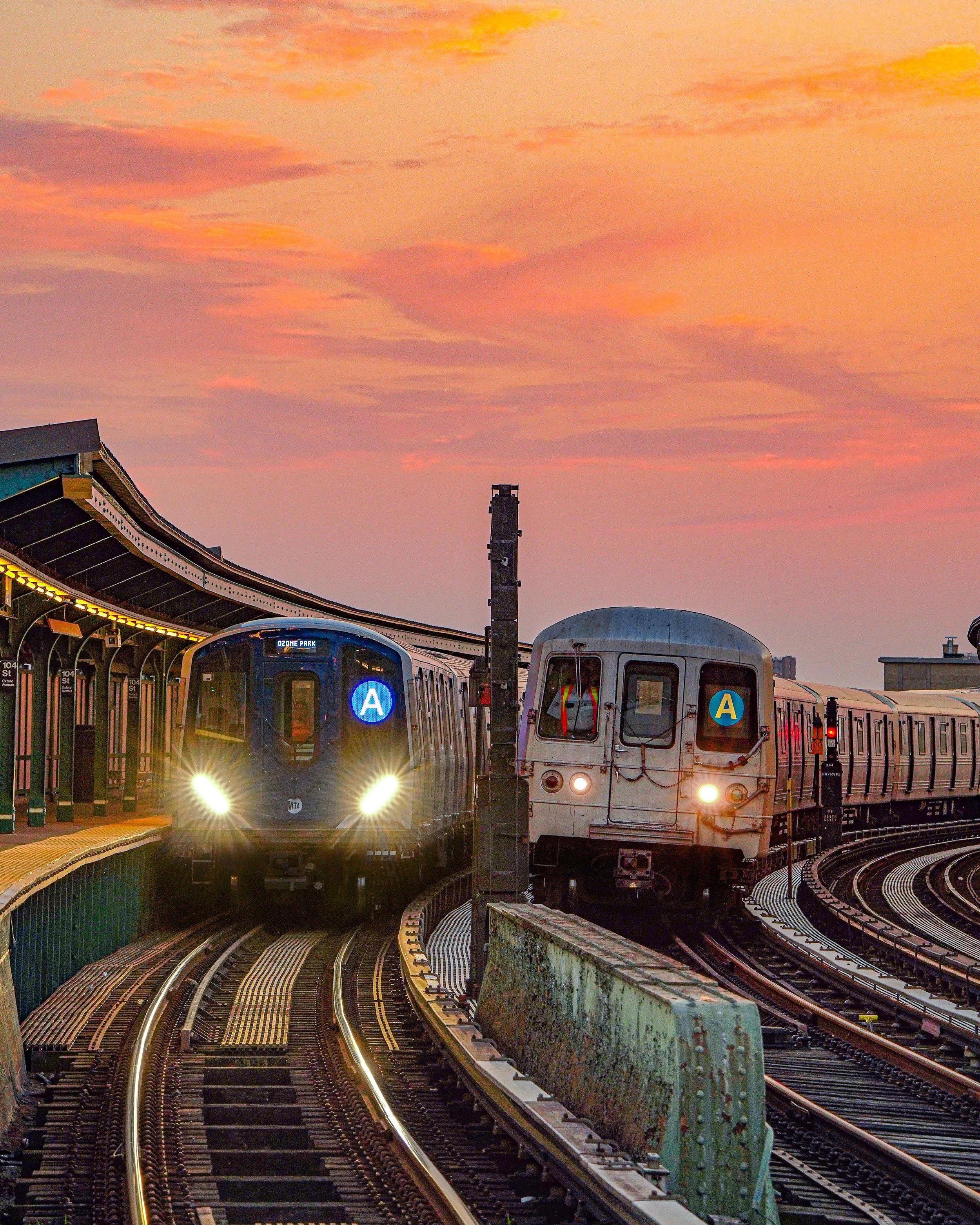 Two subway trains on an elevated track at sunset, with a colorful sky in the background.