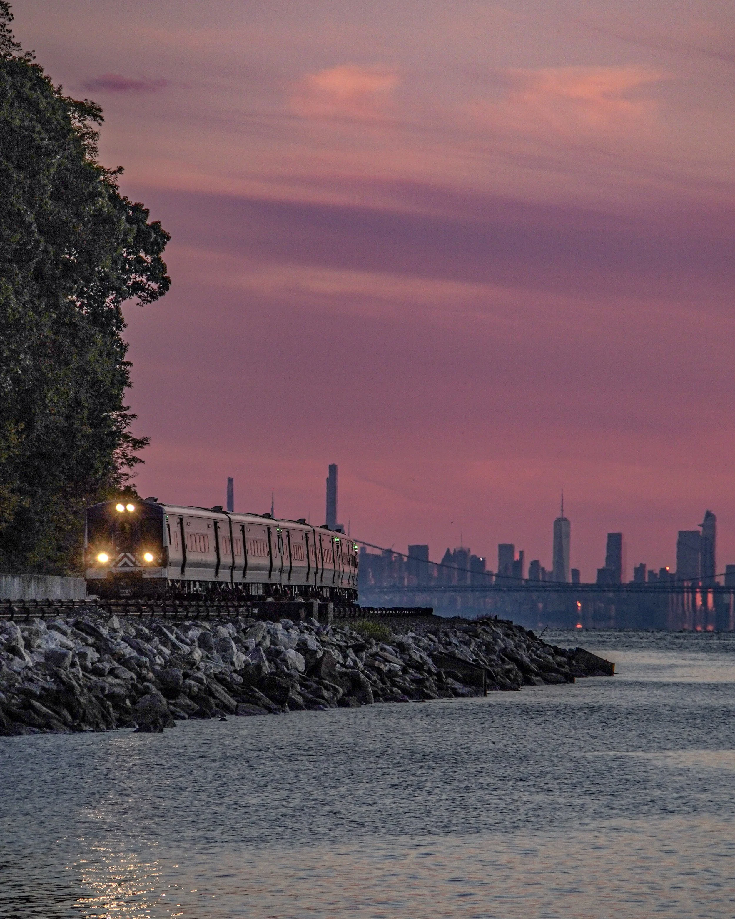 A train traveling along a rocky shoreline during sunset with a city skyline and bridge in the background.