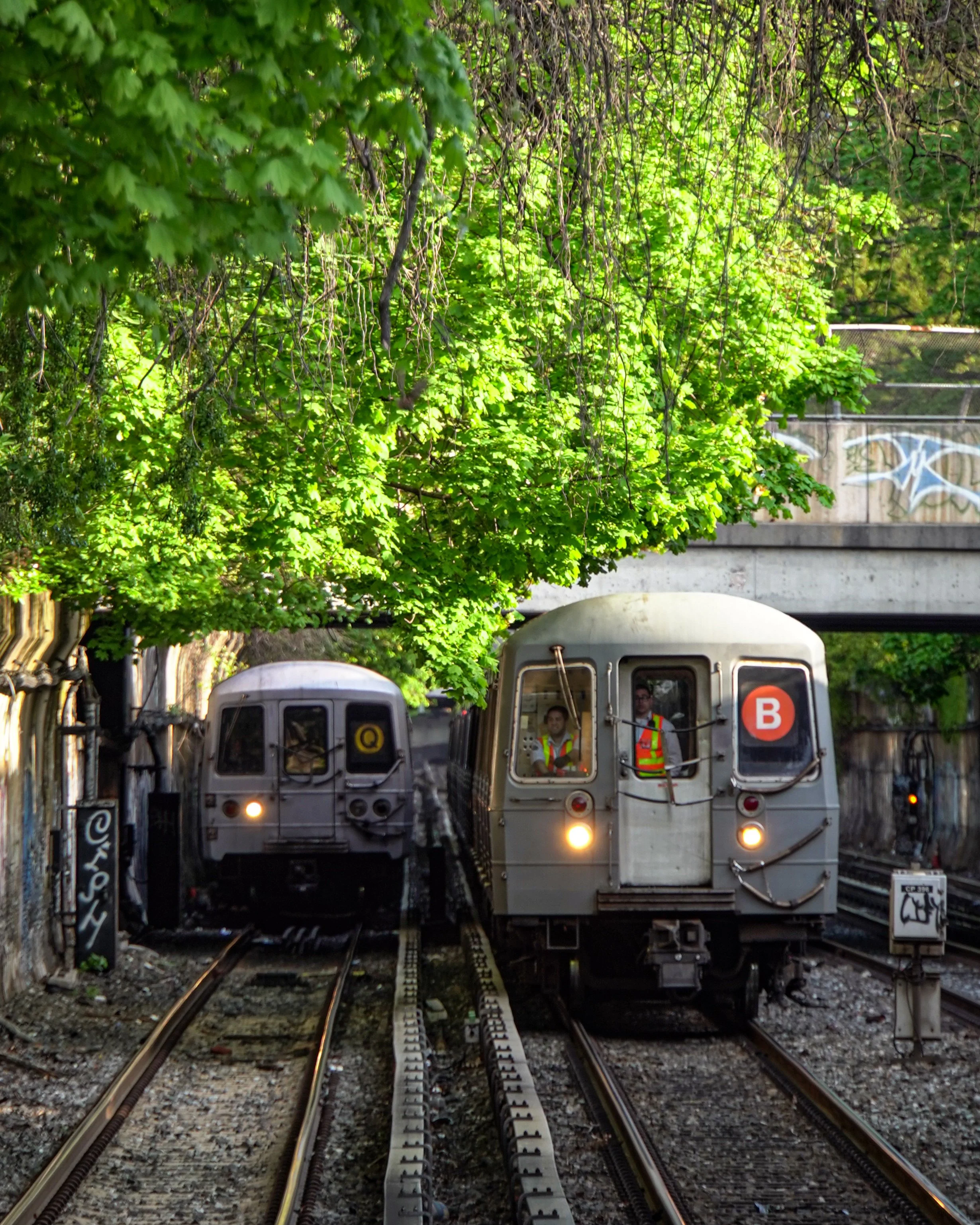 Two subway trains on parallel tracks beneath a green, leafy tree overpass, with three workers visible inside the train on the right, wearing reflective vests.