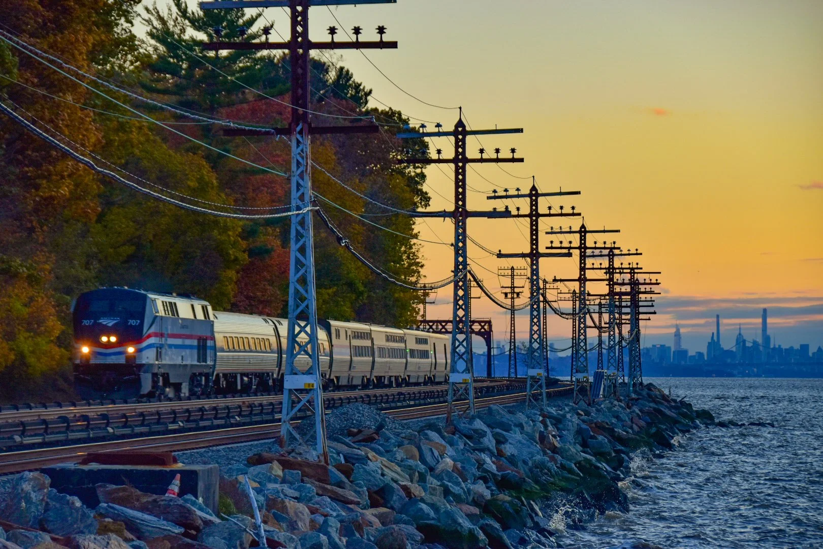 A train traveling along a railroad track beside a body of water during sunset, with a city skyline in the background and trees with autumn foliage on the left.