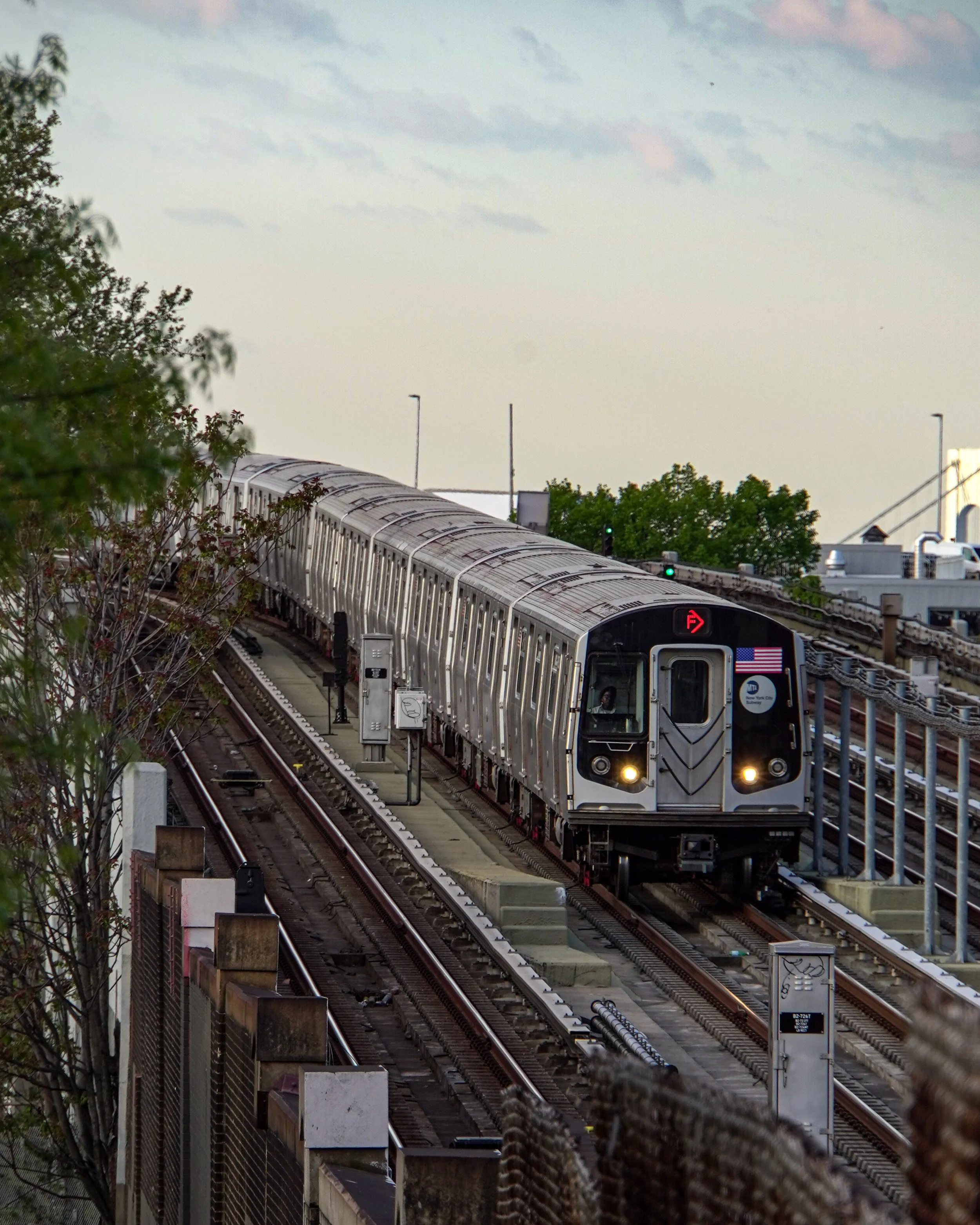 An NYC subway train on elevated tracks during daytime, with trees and a cloudy sky in the background.