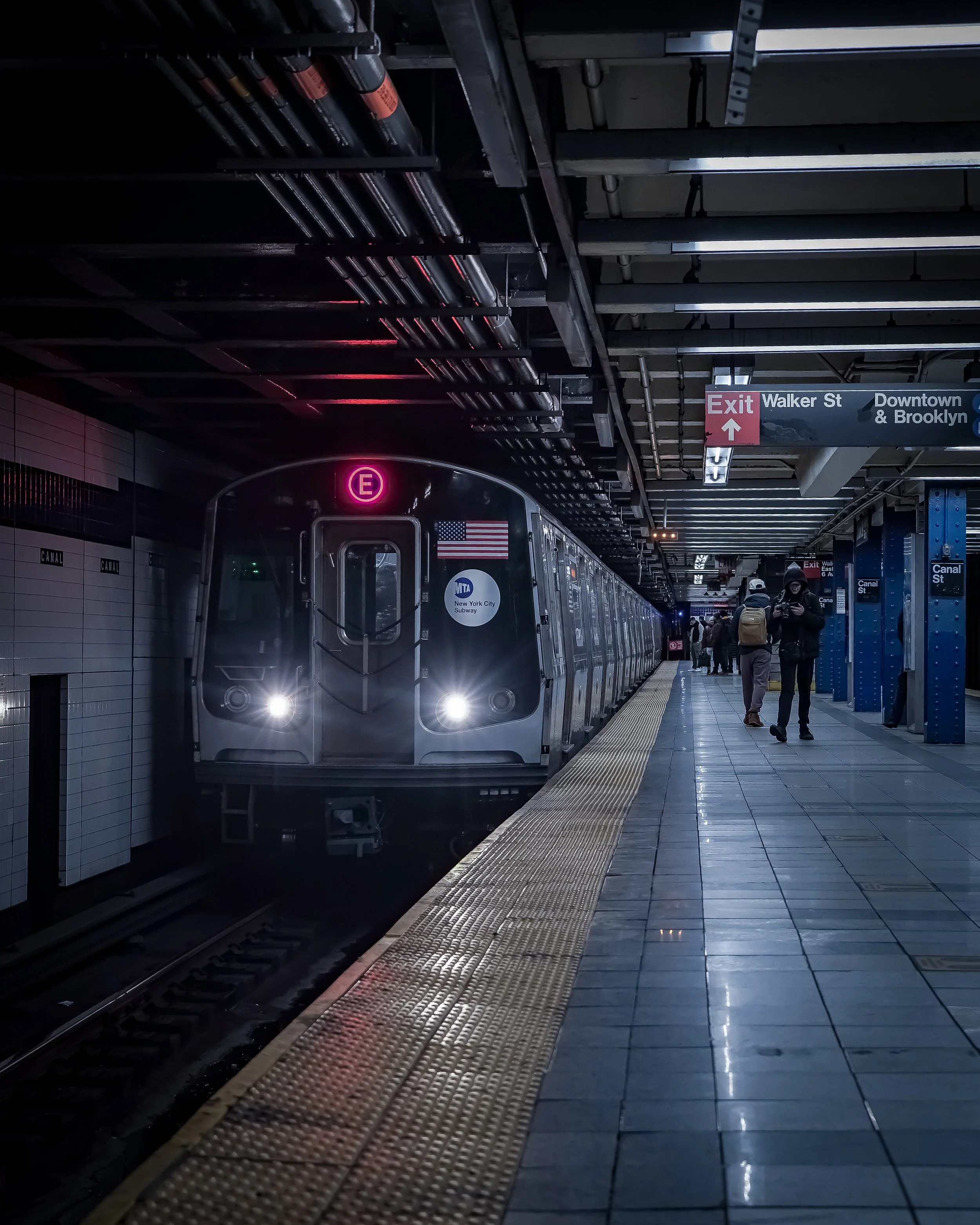 An NYC subway train at an underground station, with passengers walking on the platform. The train has illuminated headlights and an electronic sign showing the letter 'E'. The station has signs for exits and directions to various streets.