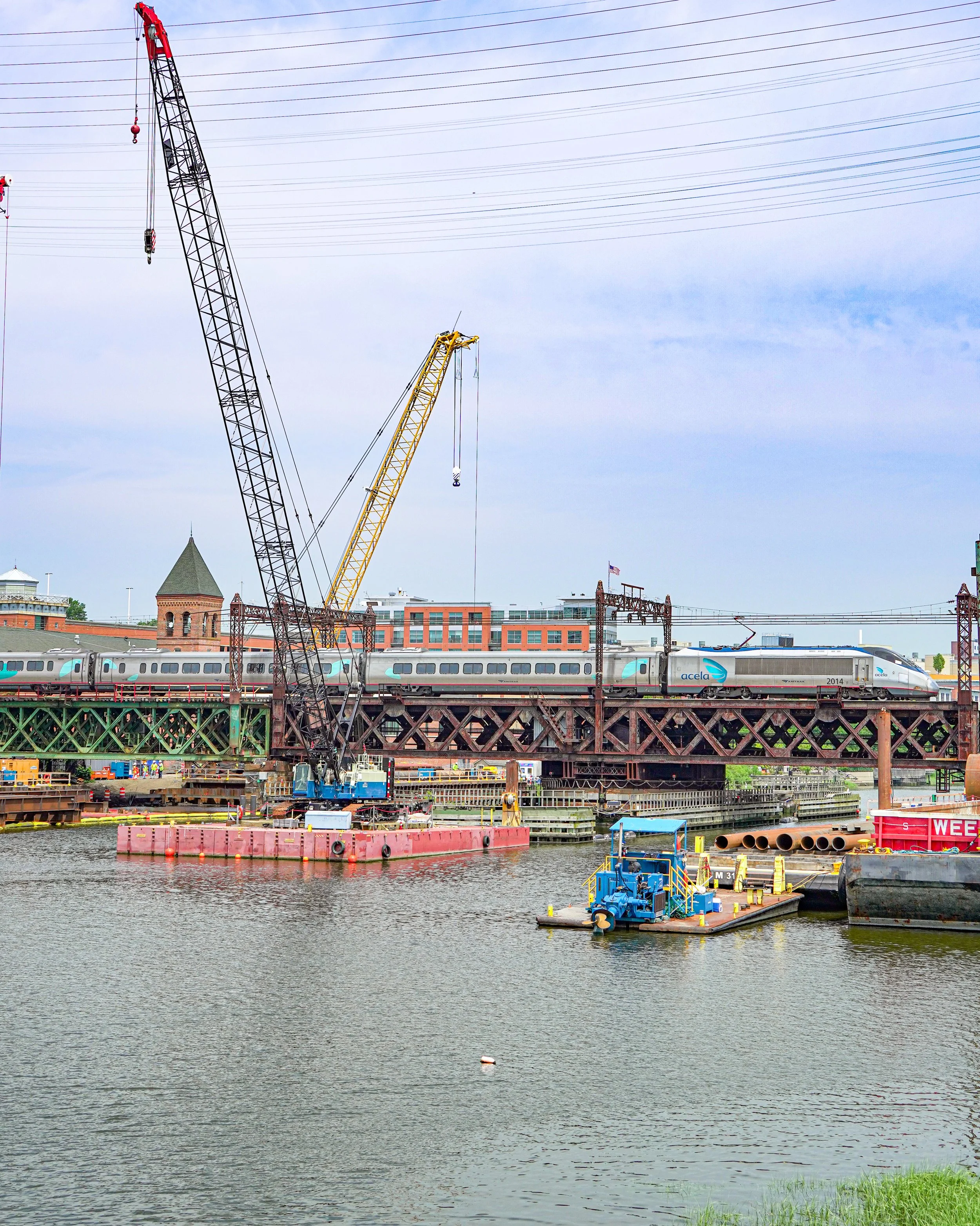 A train traveling on a bridge over a body of water, with construction cranes and buildings in the background.