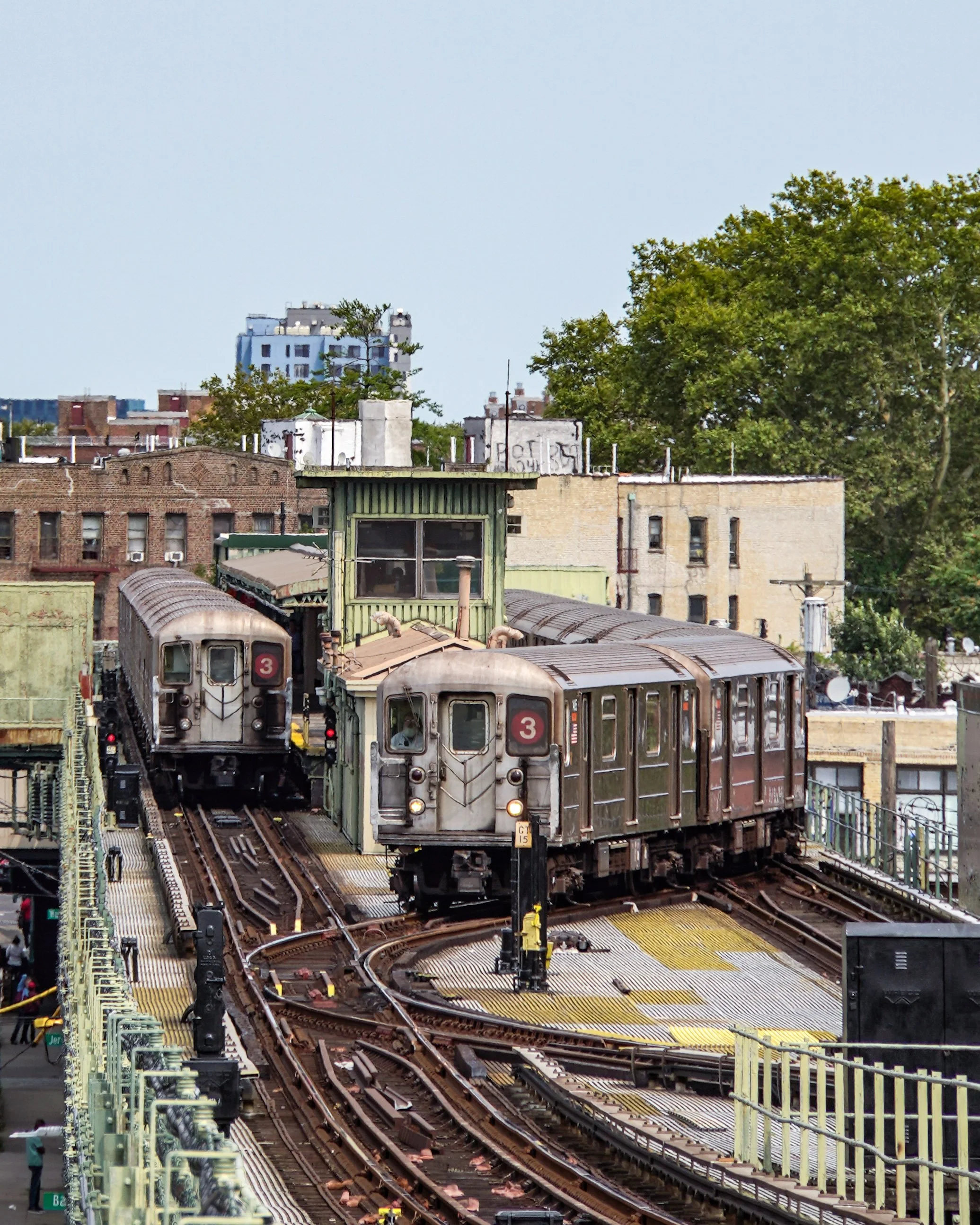 Two vintage subway trains on an elevated track, with a cityscape and green trees in the background.