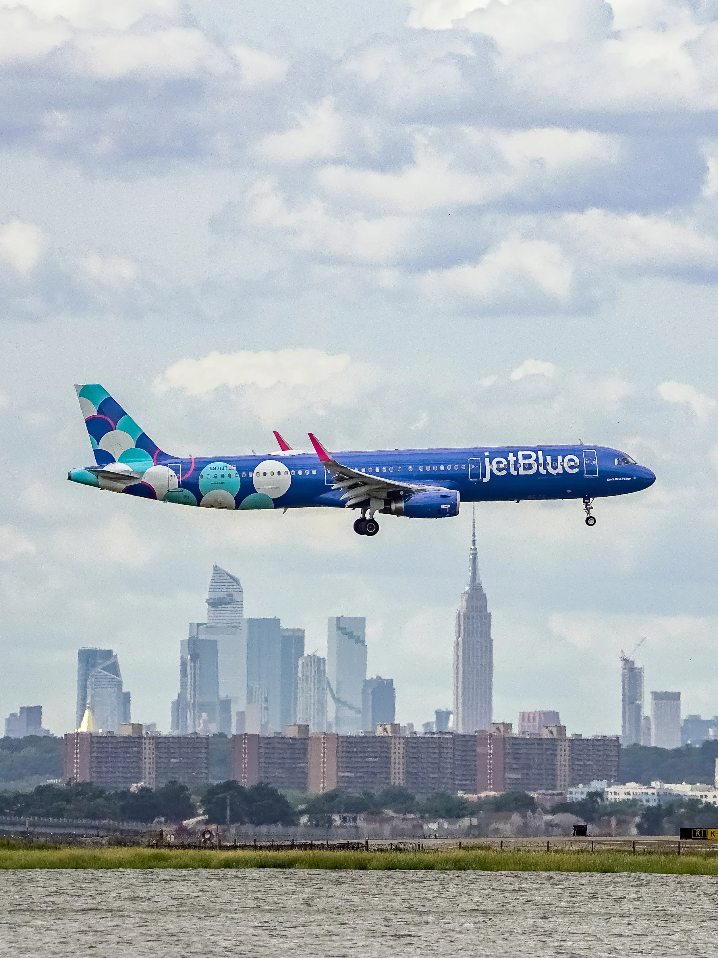 An airplane with a blue fuselage and cartoon bubble design on the tail flies over a city skyline with the Empire State Building visible in the background.