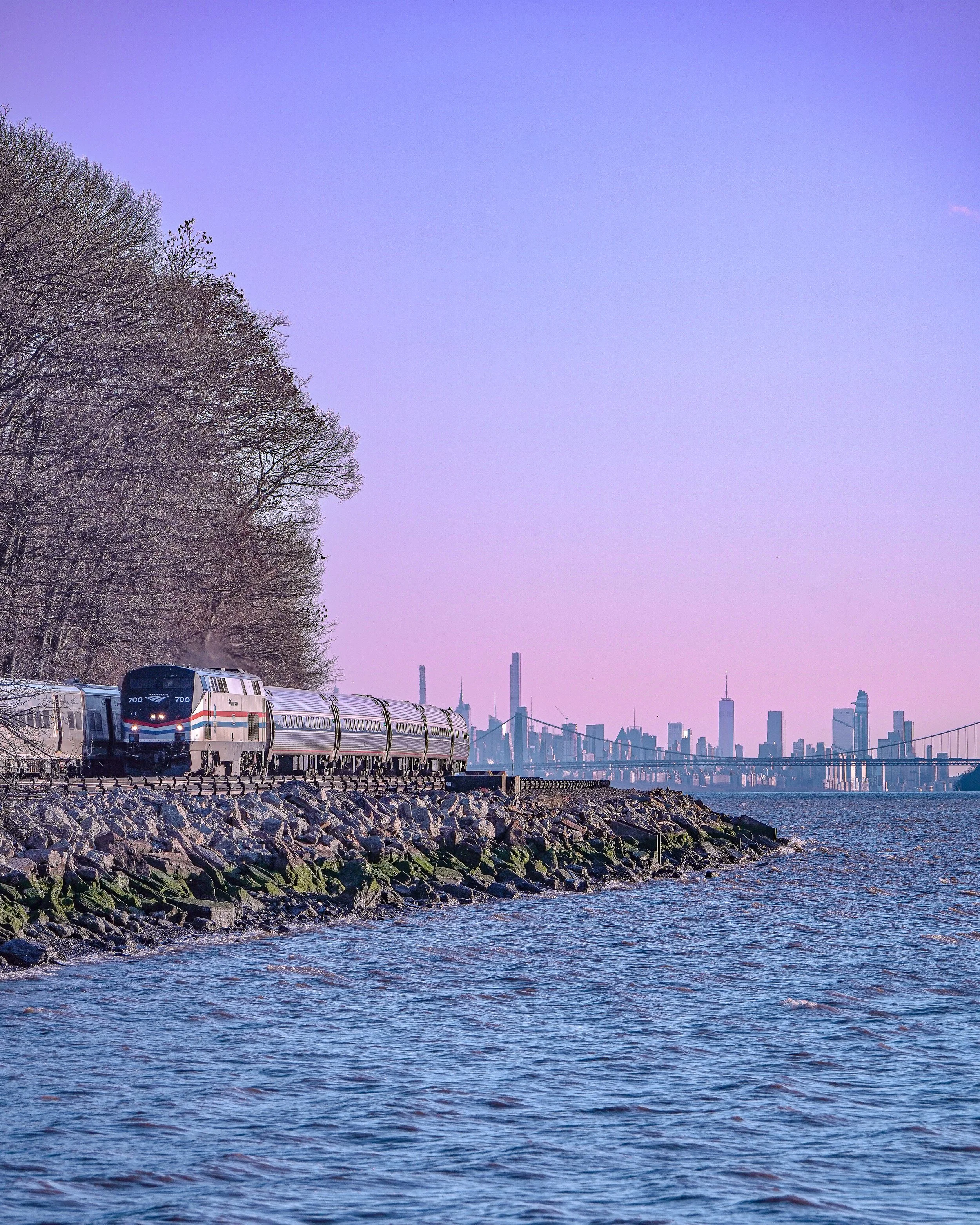 A train traveling along a rocky shoreline waters with a city skyline in the background during dusk or dawn.