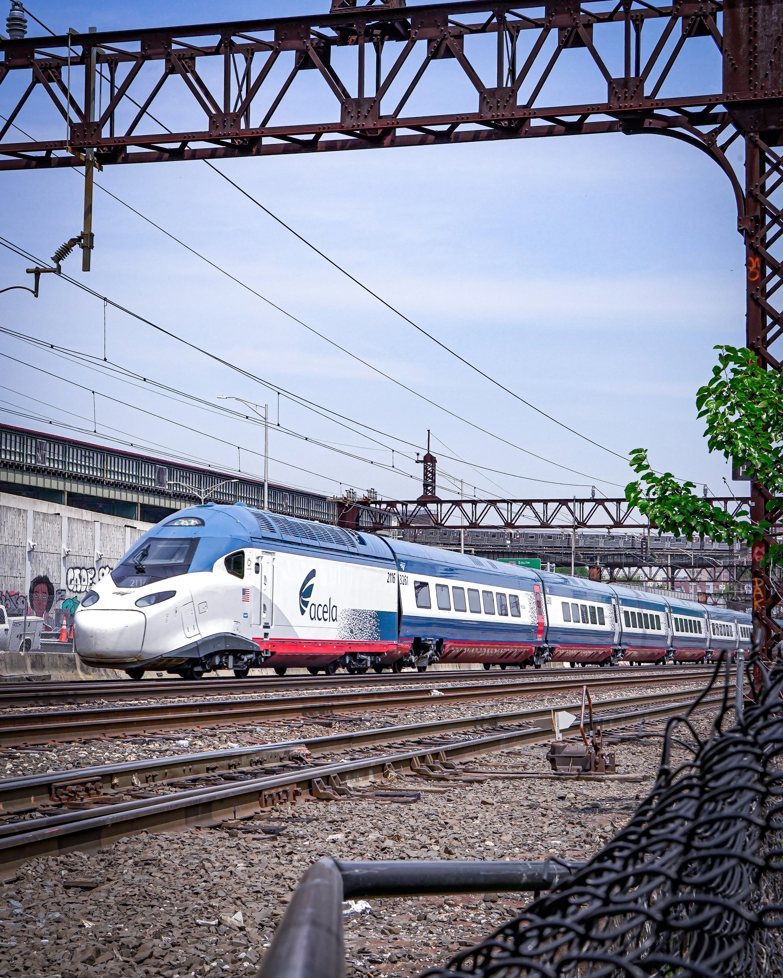 A white and blue Acela train traveling on railway tracks under a metal bridge with power lines overhead and graffiti in the background.