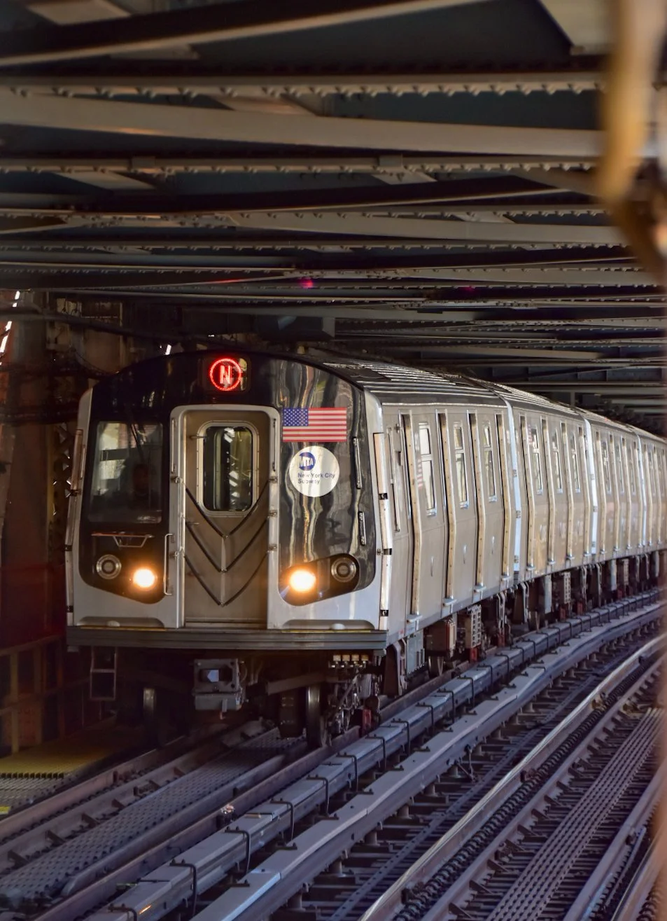 New York City subway train on elevated tracks, with headlamps on, under an urban bridge structure.