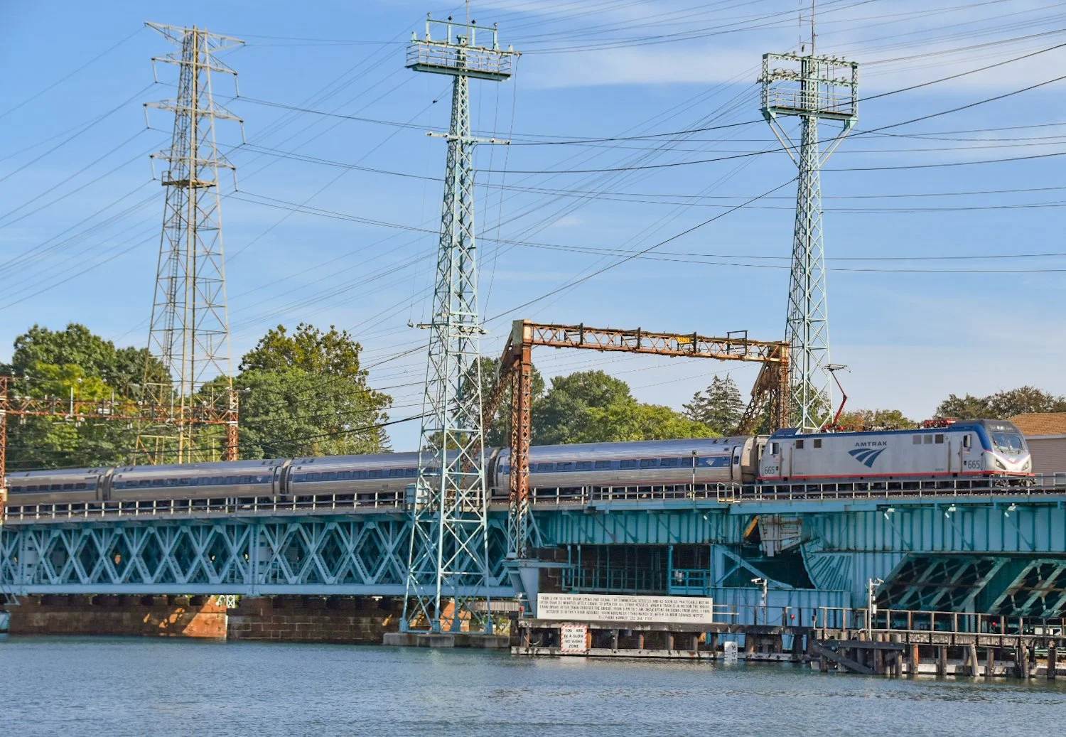 A sleek Amtrak train traveling over a bridge above a body of water, with power lines and electrical towers in the background, under a clear blue sky.