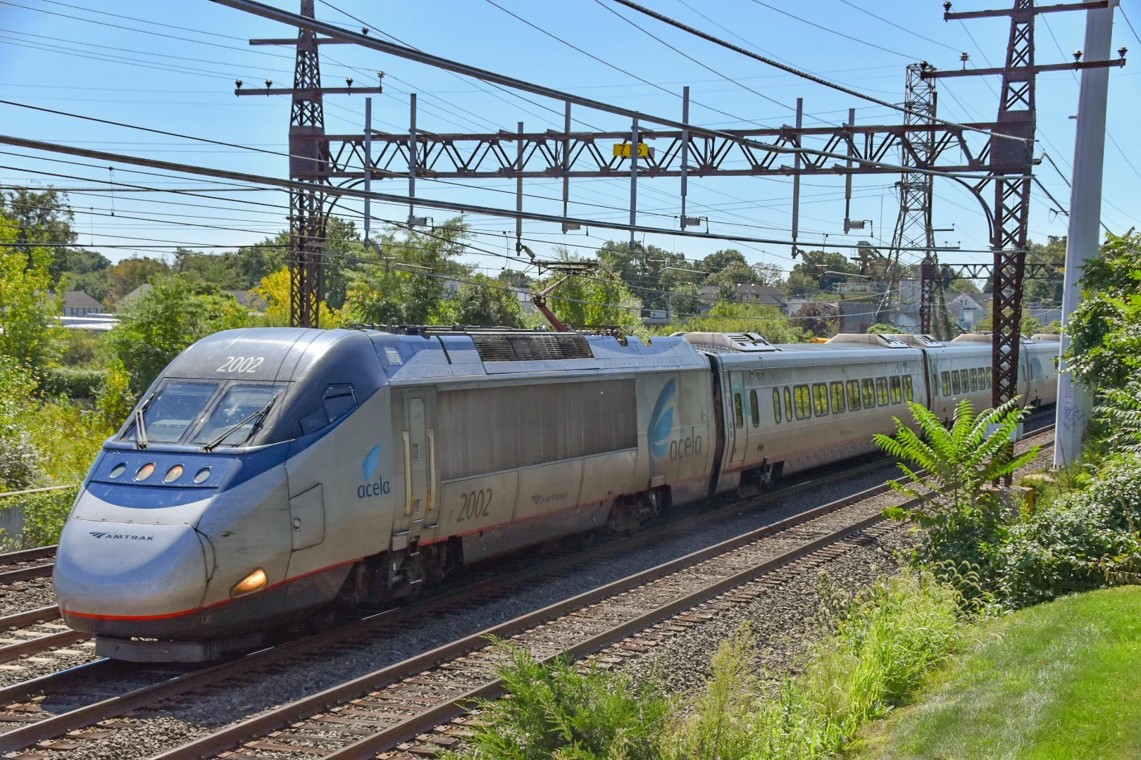 An Amtrak Acela train traveling on railroad tracks surrounded by green trees and bushes with clear blue sky overhead.