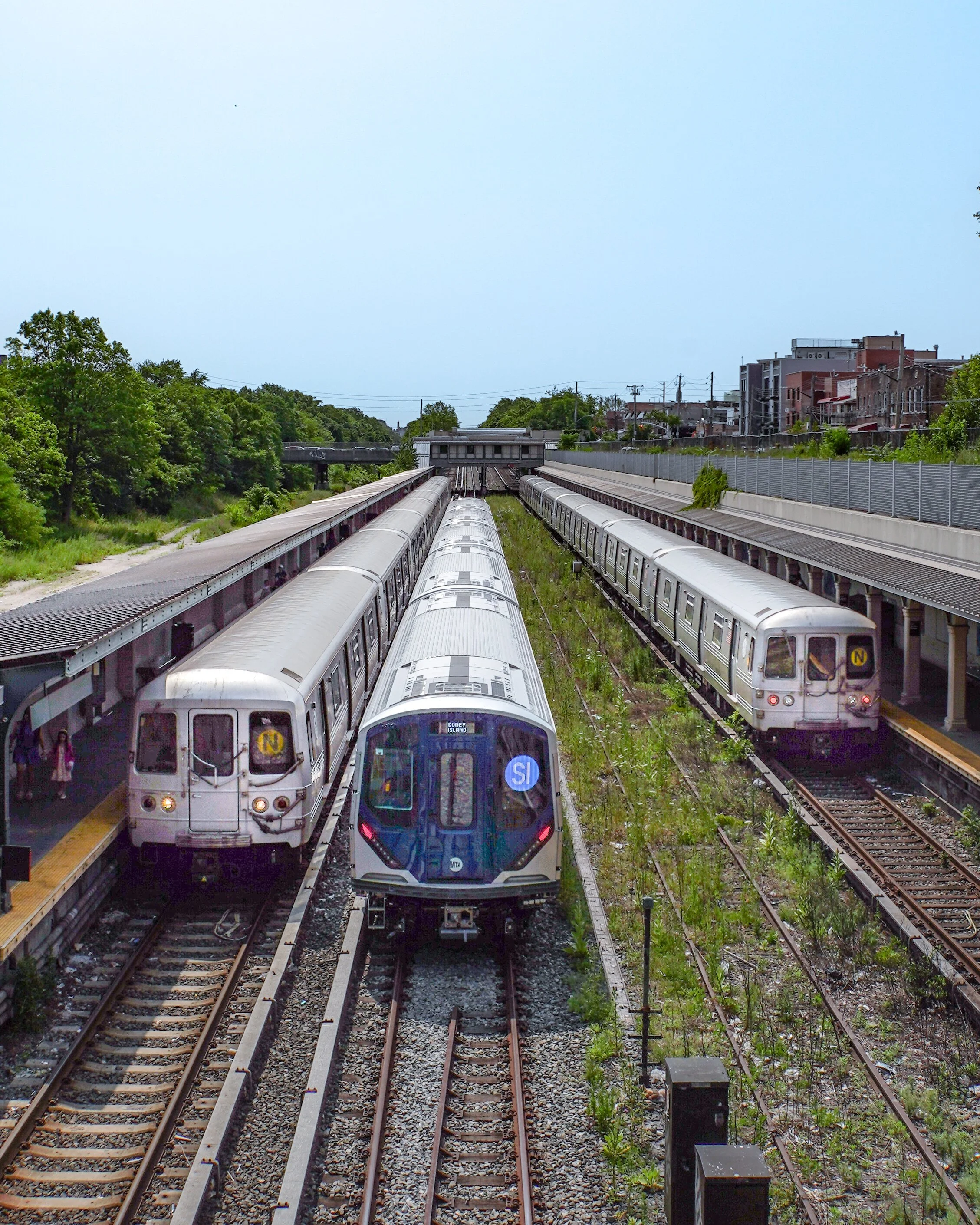 Three subway trains at a station platform with trees on the left and buildings on the right.