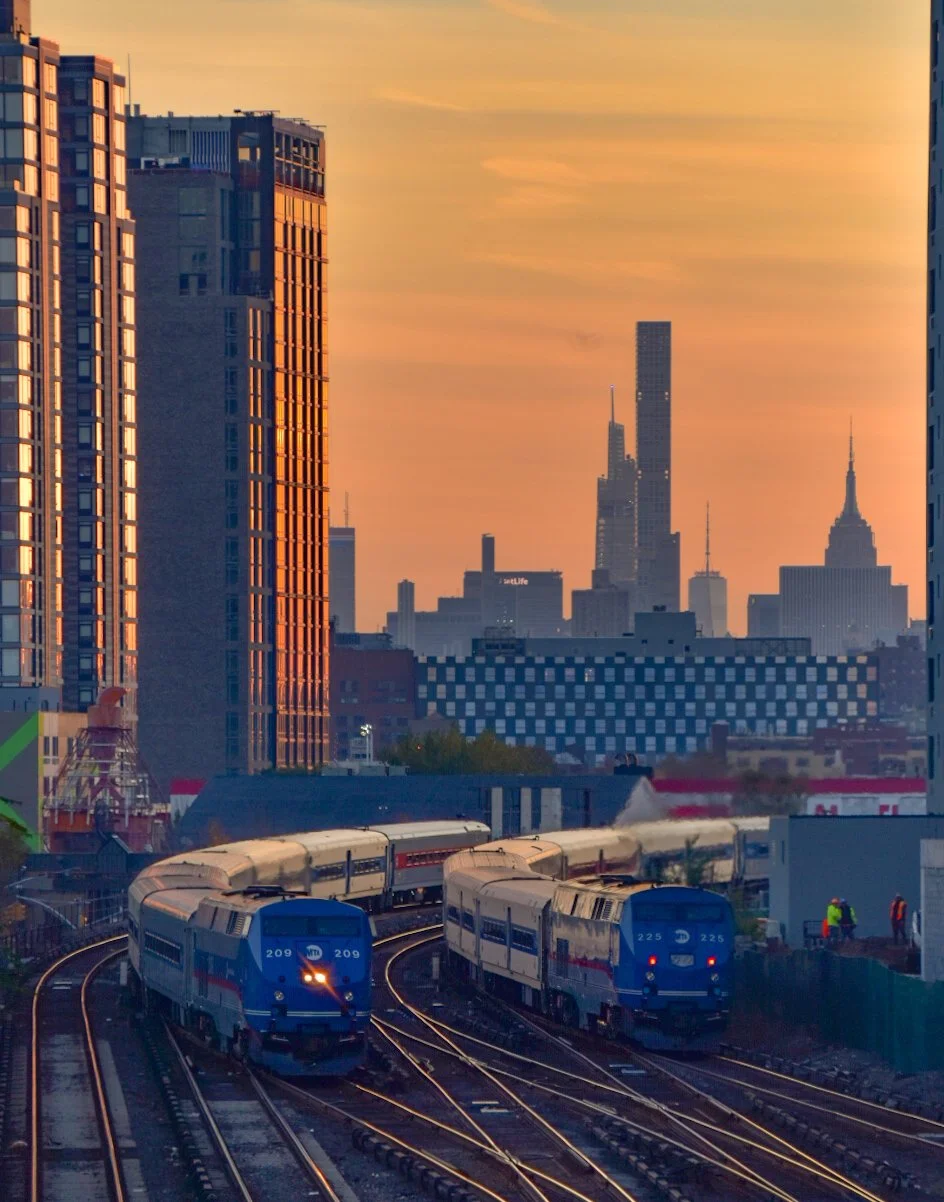 Two trains on a track with the New York City skyline in the background during sunset.