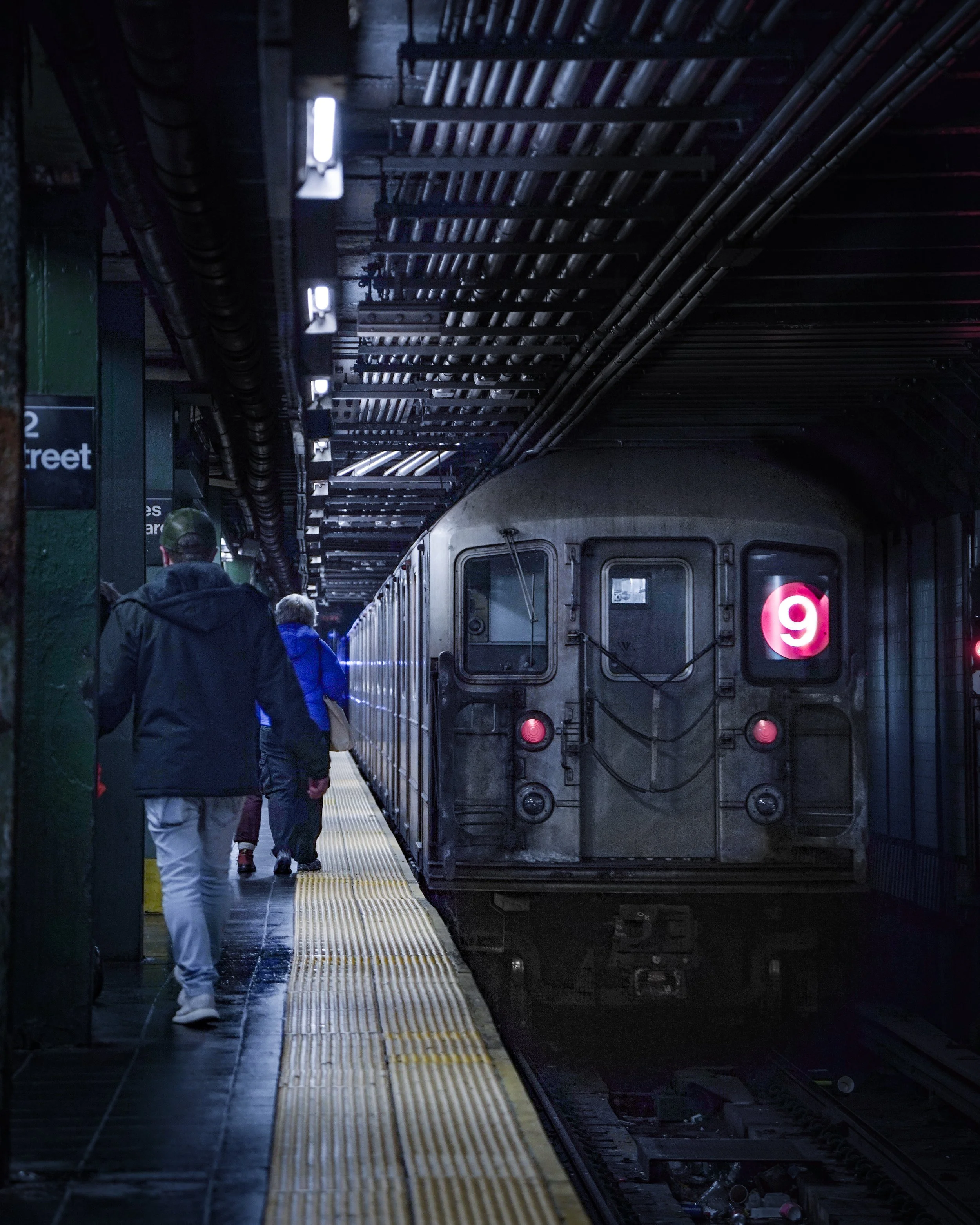 People walking in a subway station with a train departing, featuring red illuminated number 9 on the front.