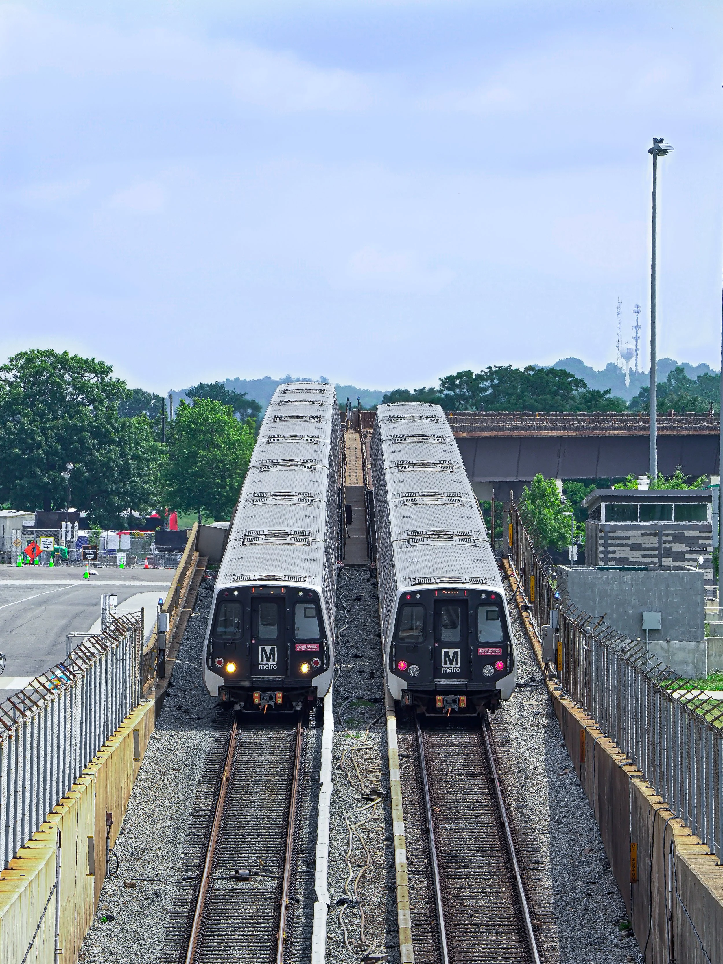 Two metro trains on parallel tracks in a station with safety fencing, trees, and a cloudy sky in the background.