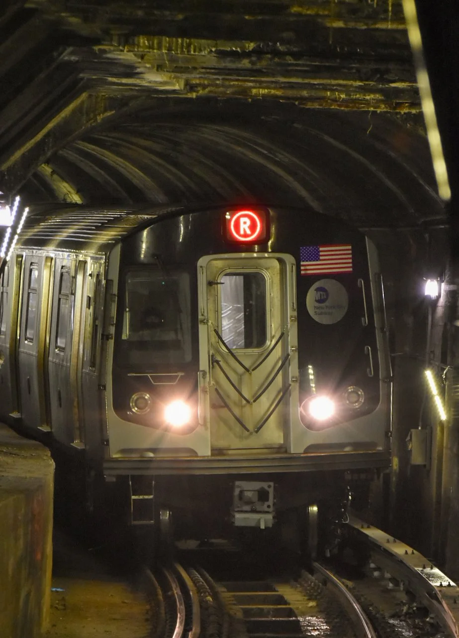 An NYC subway train on a dark tunnel track with its headlights on, displaying an illuminated red circle with the letter 'R' indicating the route, and American flag and NYC transit stickers on the front.