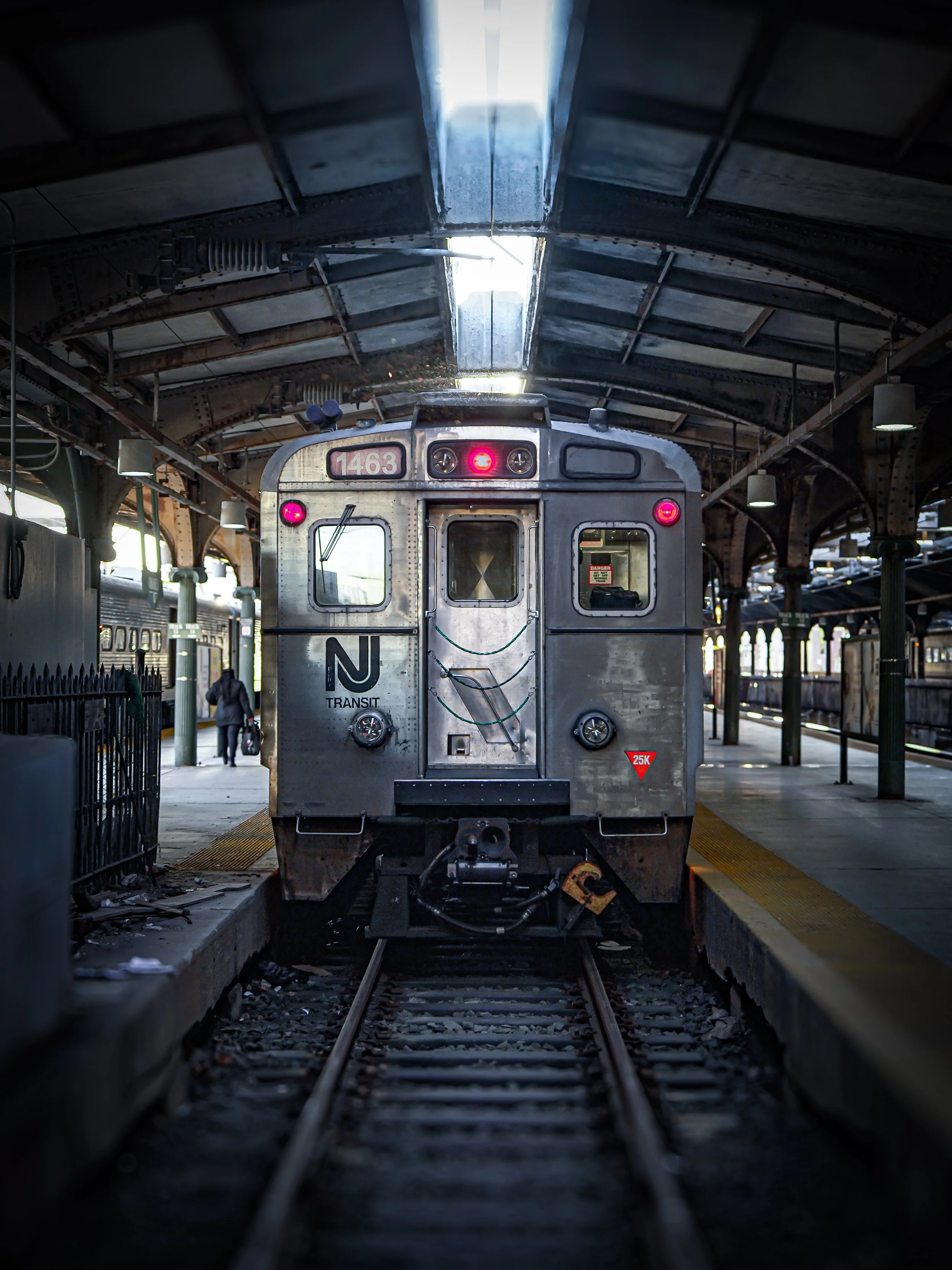 Front view of a silver subway train with pink lights inside a station, with tracks in the foreground and platform on the sides.