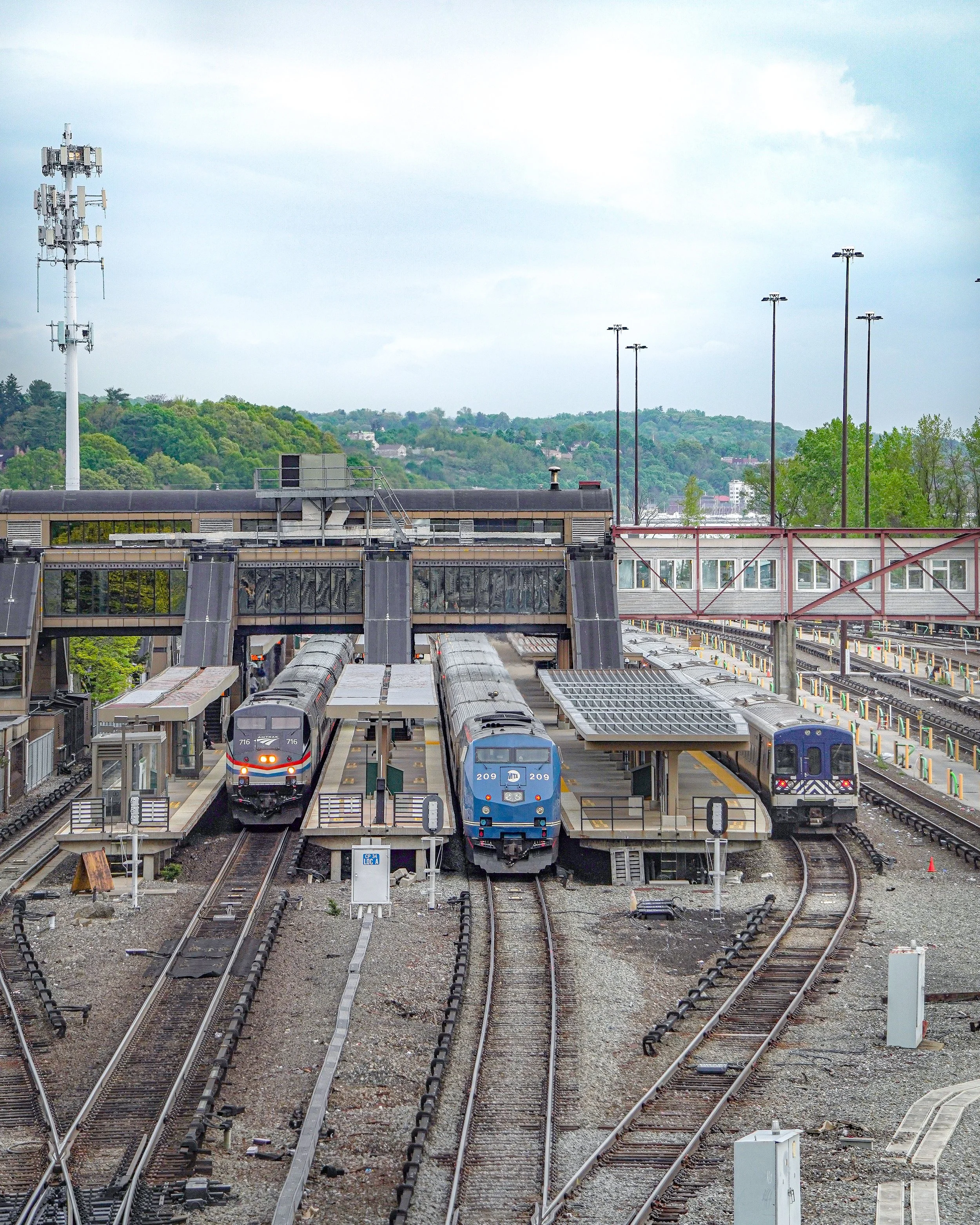 Three trains at a train station with overhead structures, in an urban area with green trees and hills in the background.